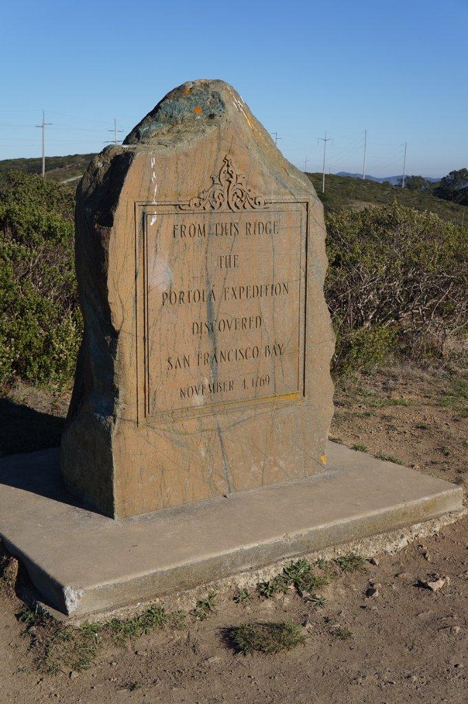 Large stone monument with engraved text on a sunny, grassy hilltop.