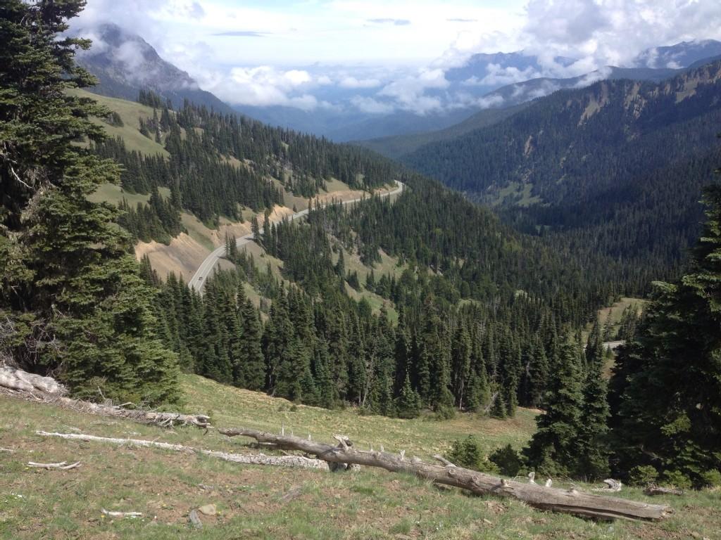 A winding road cuts through a vast, forested mountain valley under clouds.