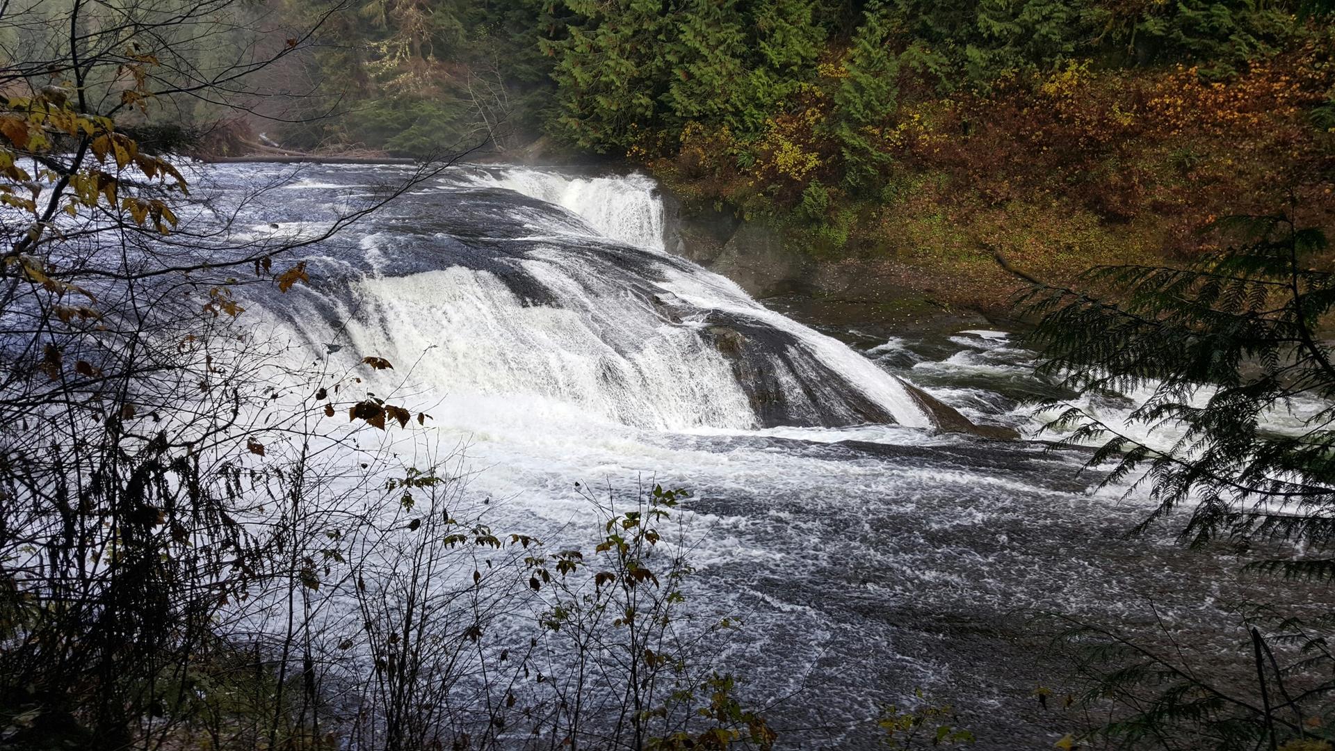 Wide, cascading waterfall over dark rocks, surrounded by autumn trees.