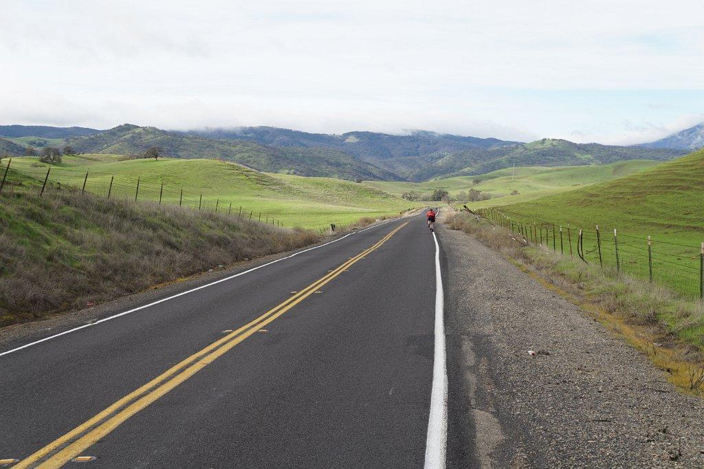 Long road through lush green hills under an overcast sky, person walking.