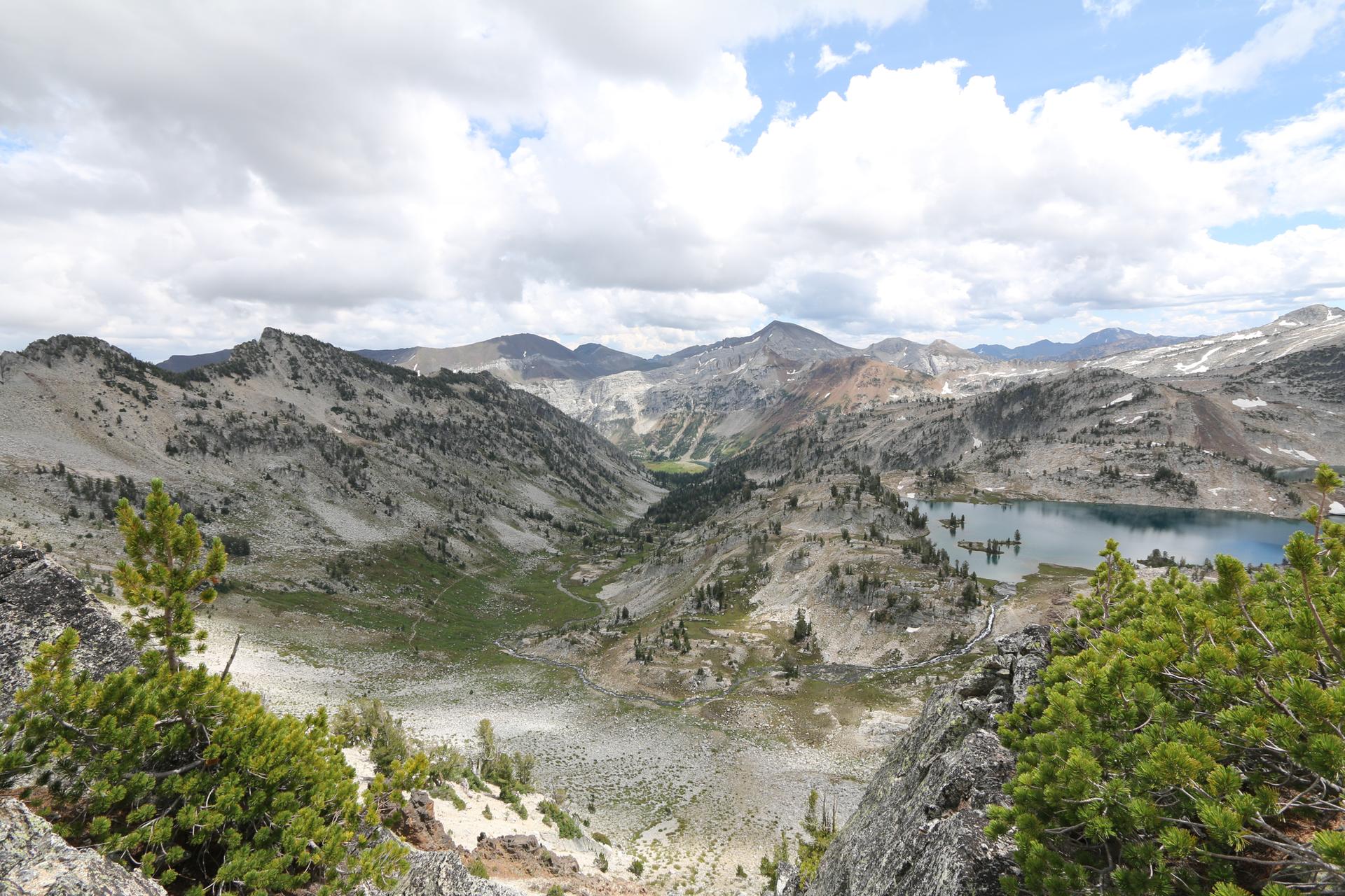Expansive mountain landscape with a valley, distant lake, and cloudy sky.