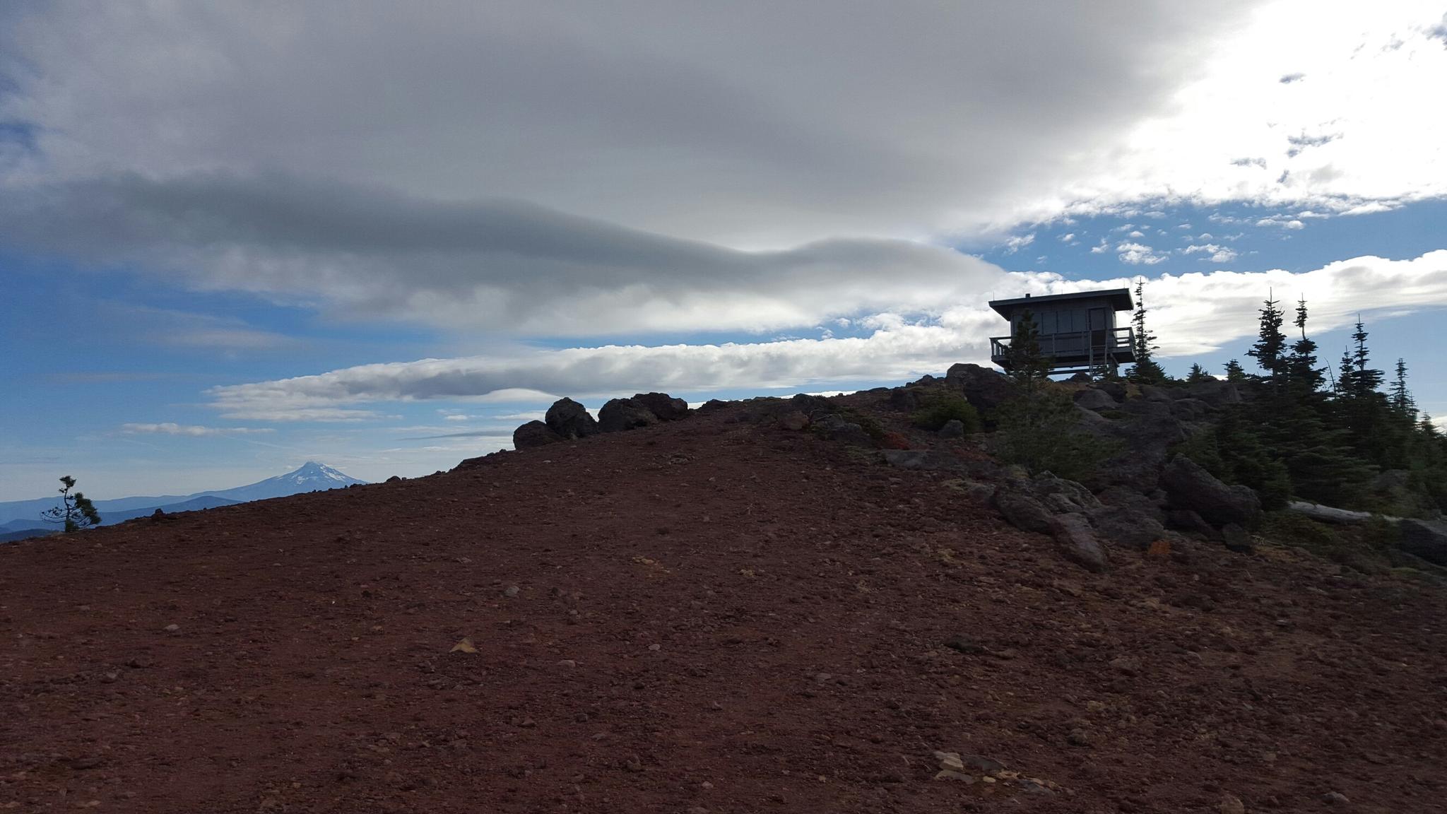 A person sits on a rocky hill near a lookout building under cloudy skies.