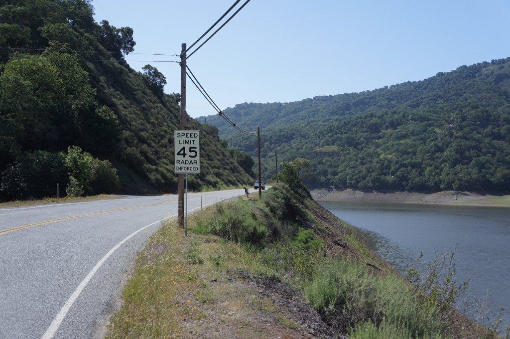 A winding road with a speed limit sign runs alongside a large body of water.