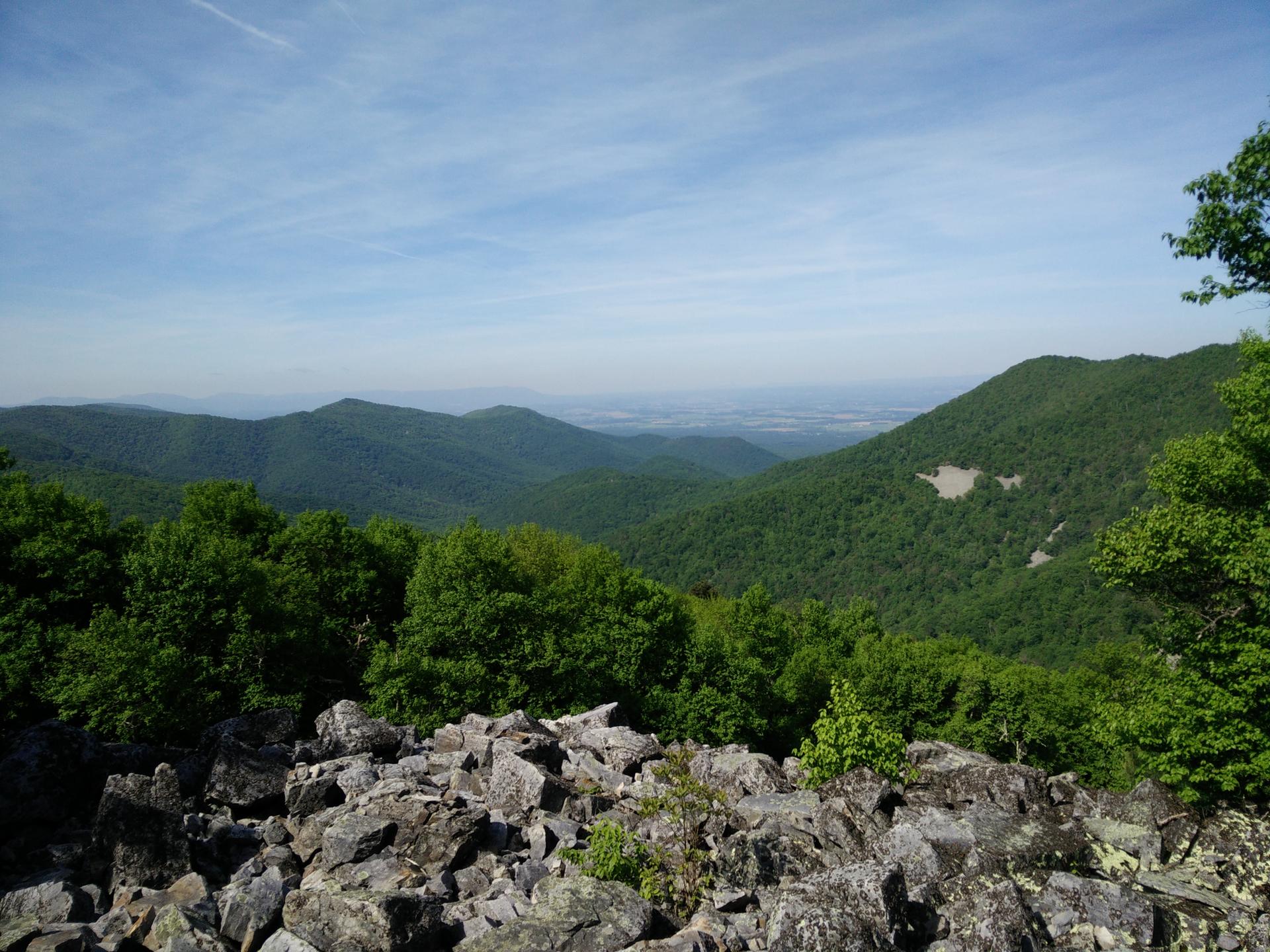 Expansive view of green forested mountains with a rocky foreground on a clear day.