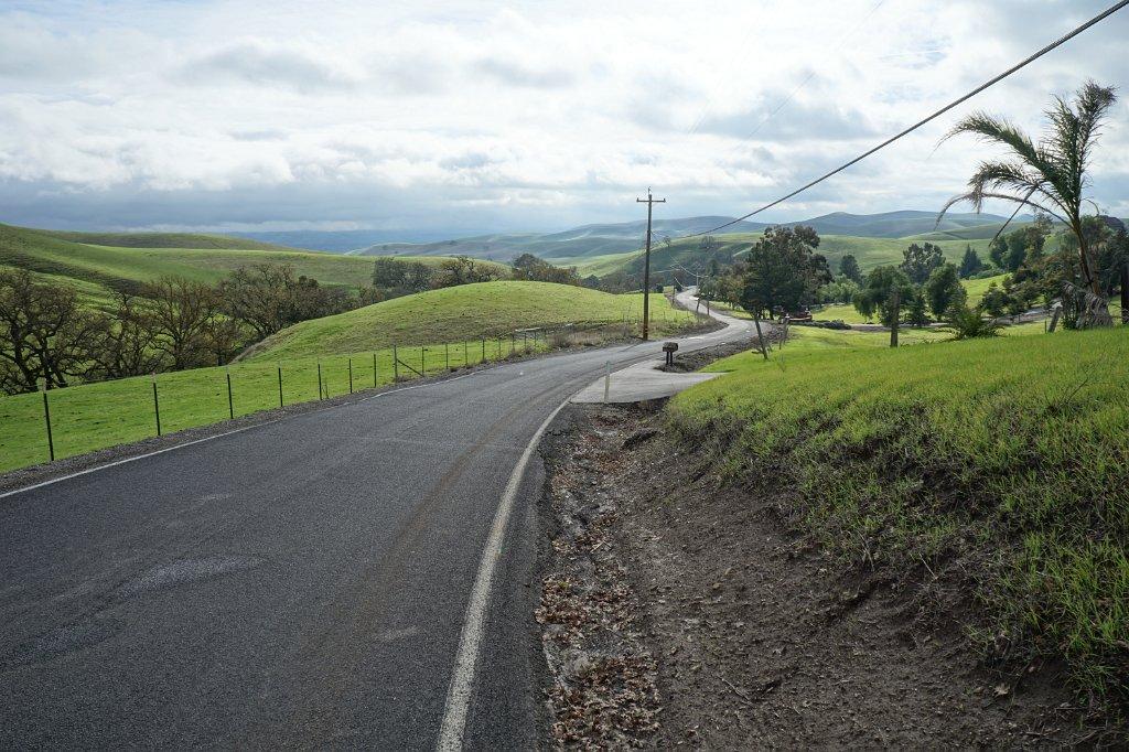 Curved paved road through green hills with scattered trees under a cloudy sky.