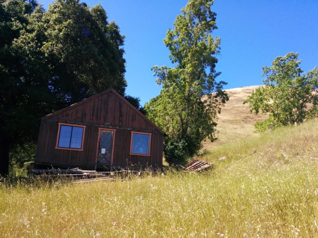 Weathered wooden cabin on a grassy hillside under a bright blue sky.