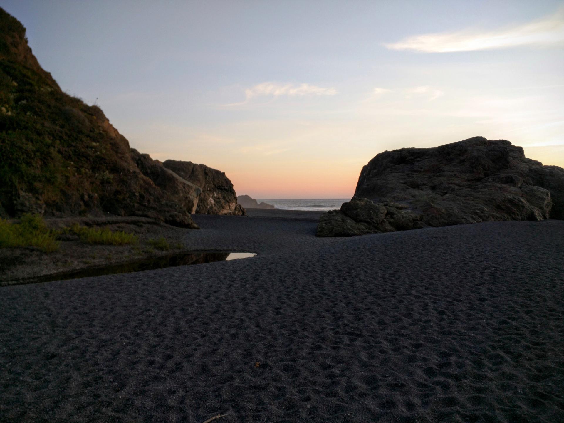 Dark sandy beach at sunset, with large rocks and distant ocean.