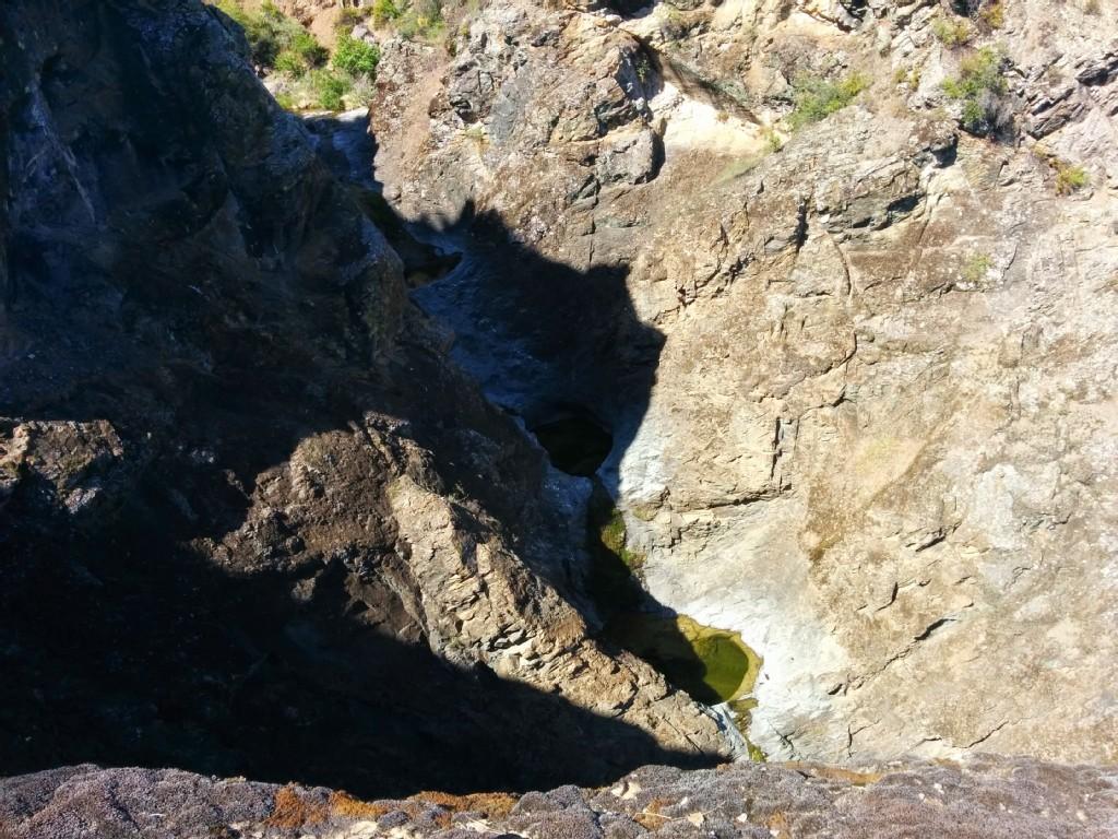 Deep rocky canyon with a shadowed pool of water below, bright sunlight.