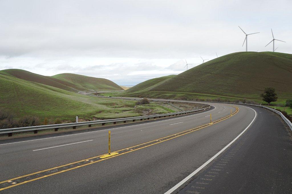 Winding road through green rolling hills under an overcast sky, with wind turbines.