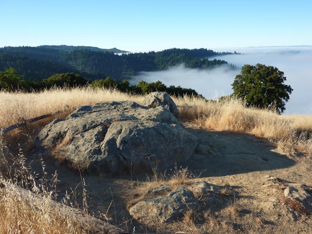 Large rock on a grassy hill overlooking a foggy valley under clear skies.