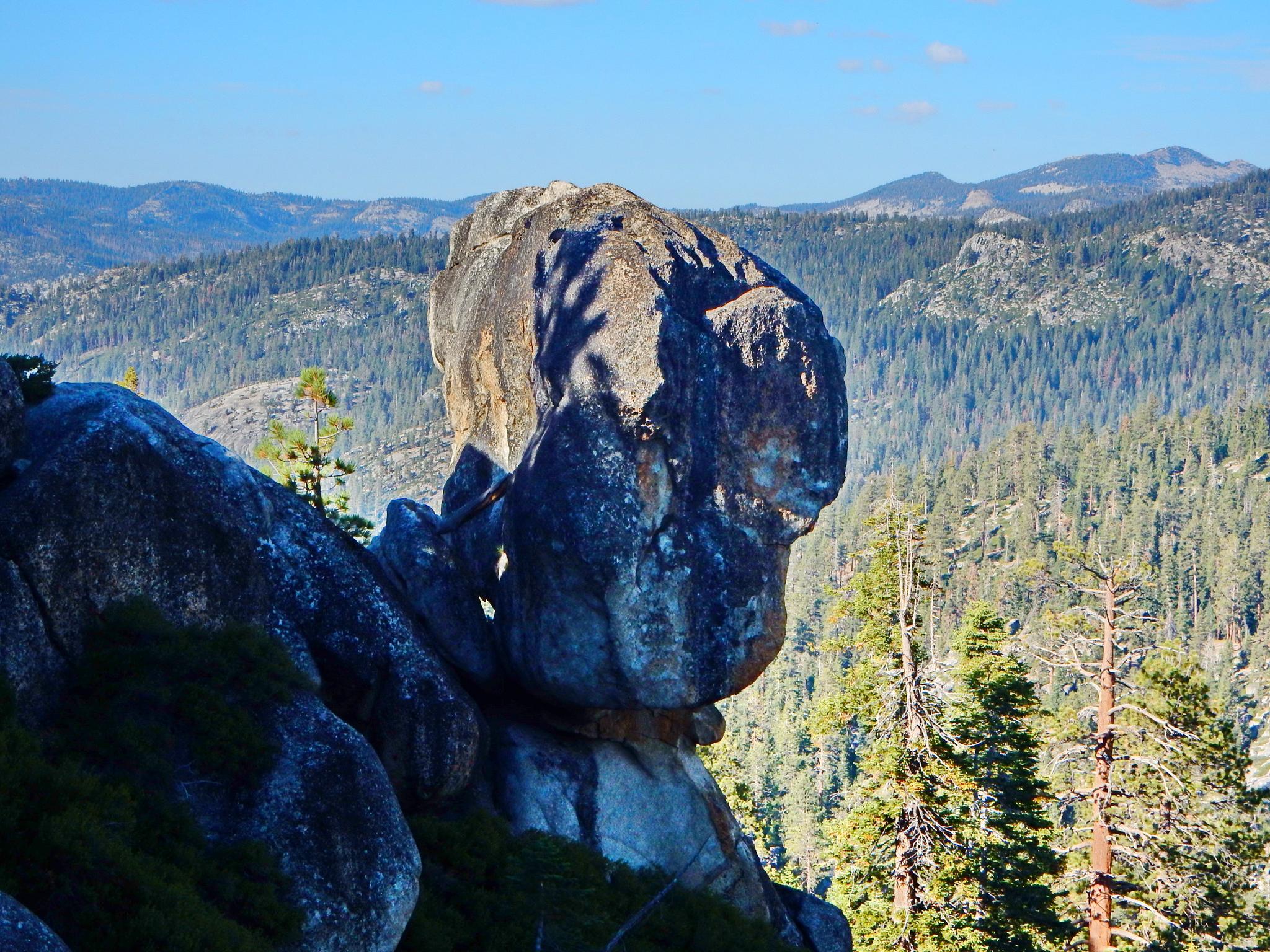 A large, weathered rock formation overlooks a vast, sunlit forested valley.