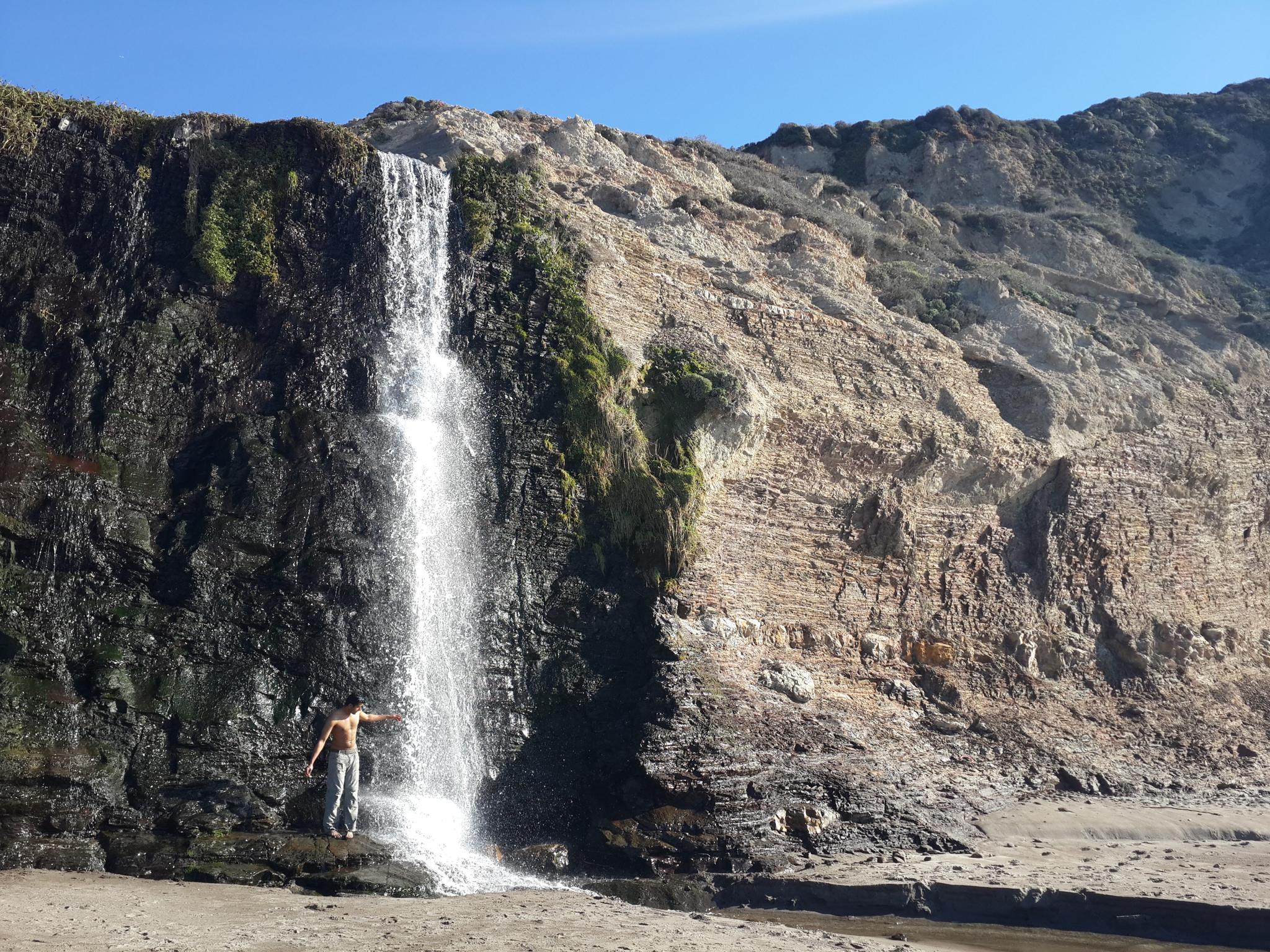 A person stands under a waterfall cascading down a rocky cliff onto a beach.