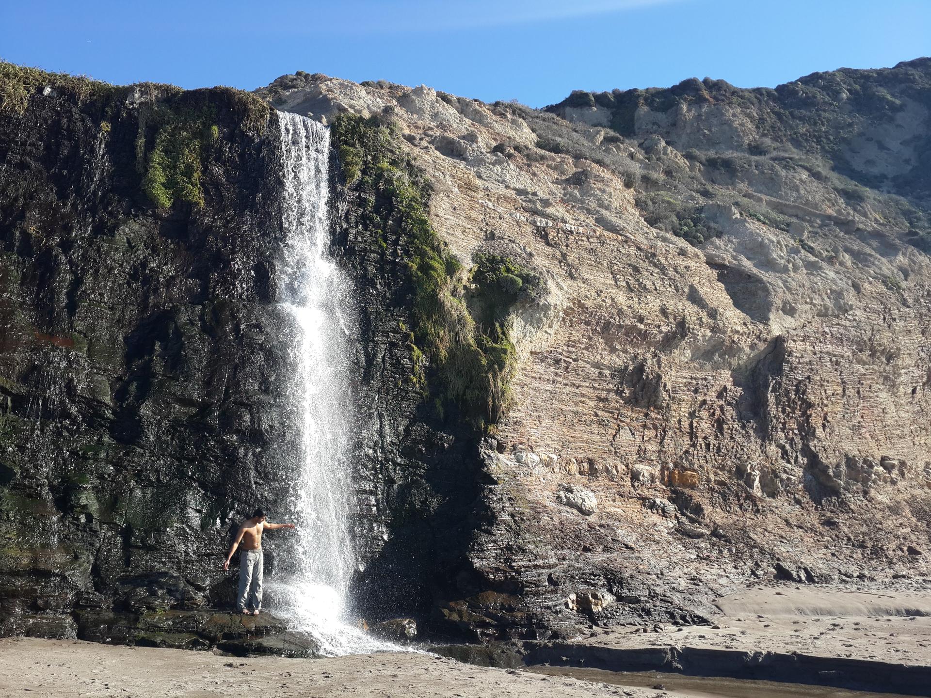 A person stands under a waterfall cascading down a rocky cliff onto a beach.