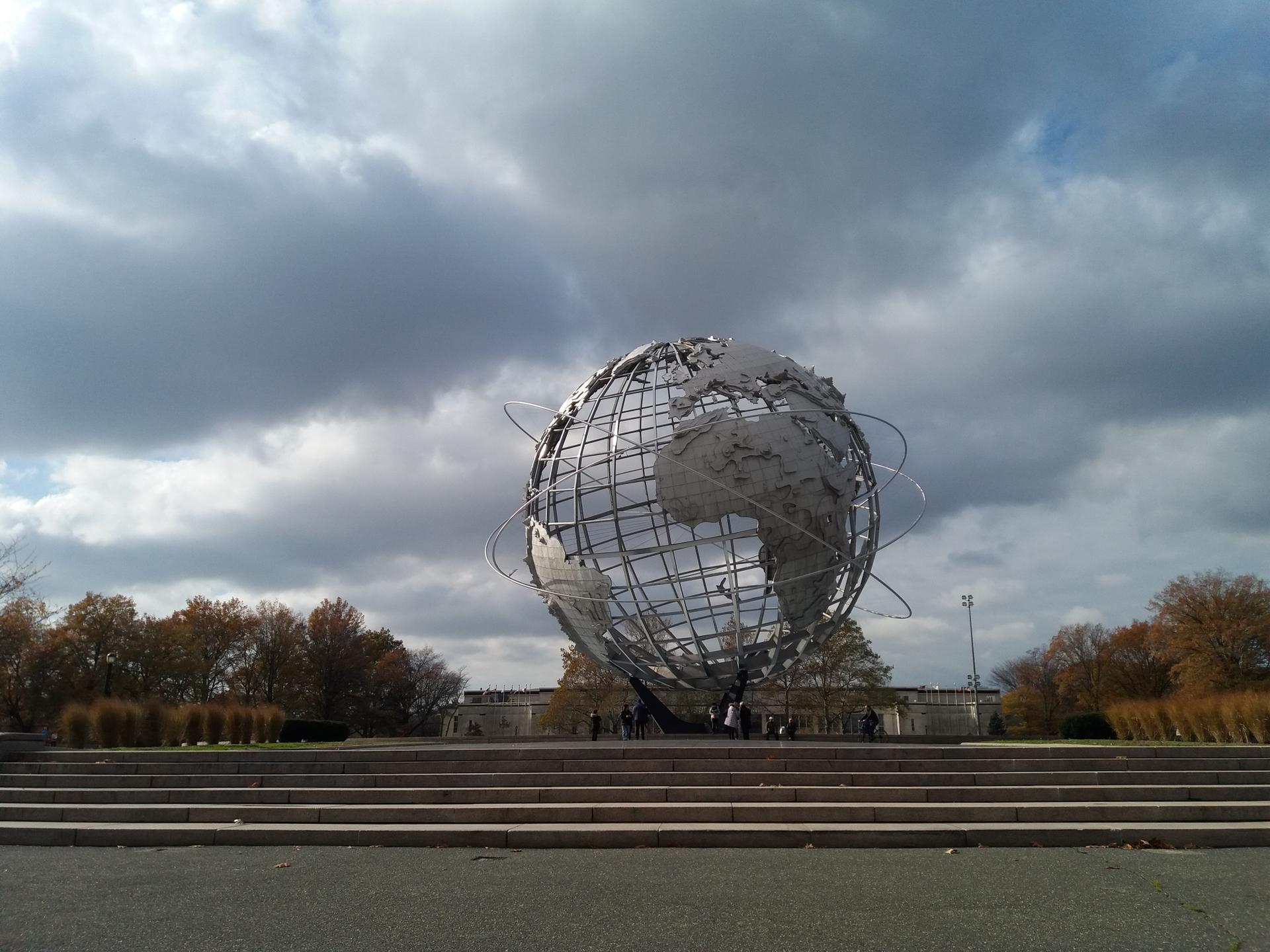 Large metallic globe sculpture viewed from steps under a dramatic, cloudy sky.