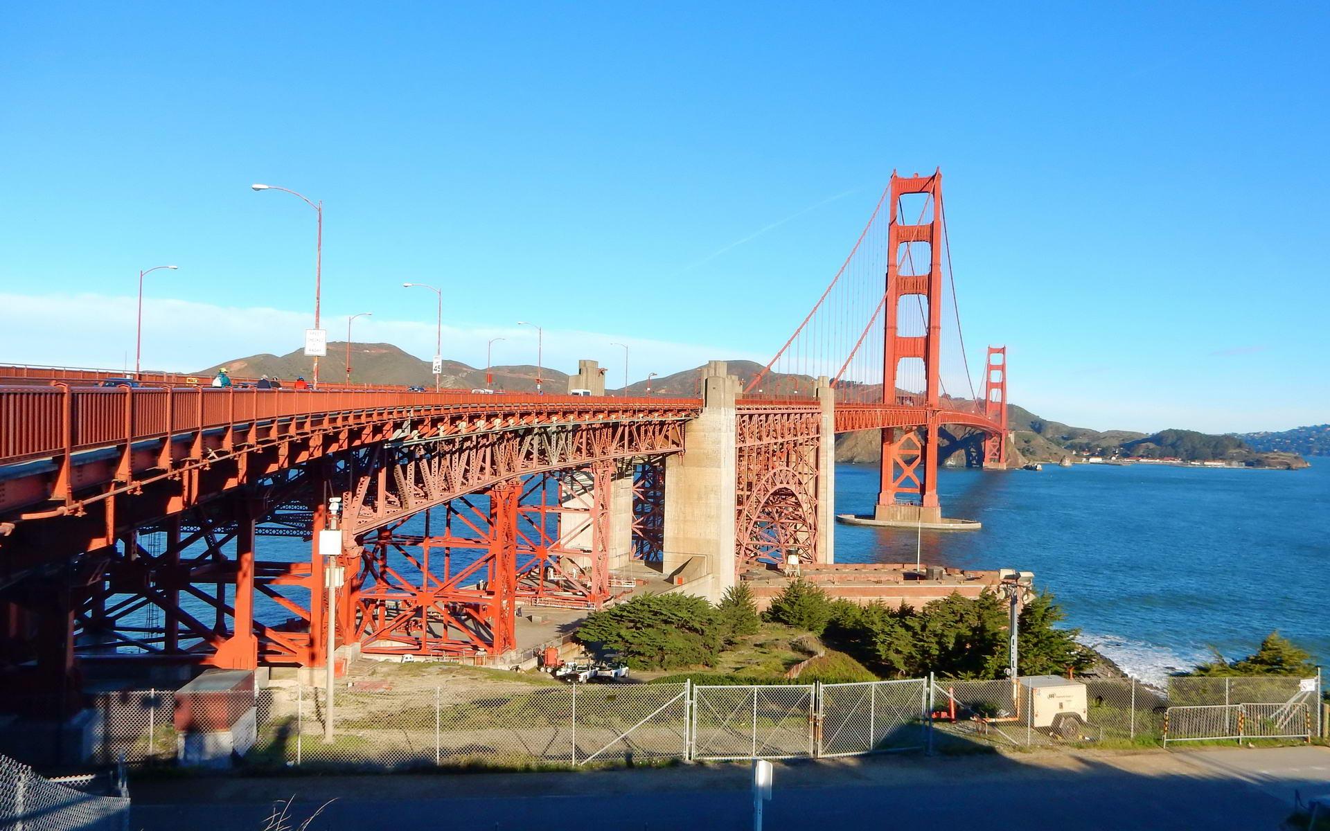 A large red suspension bridge spans blue water on a clear, sunny day.