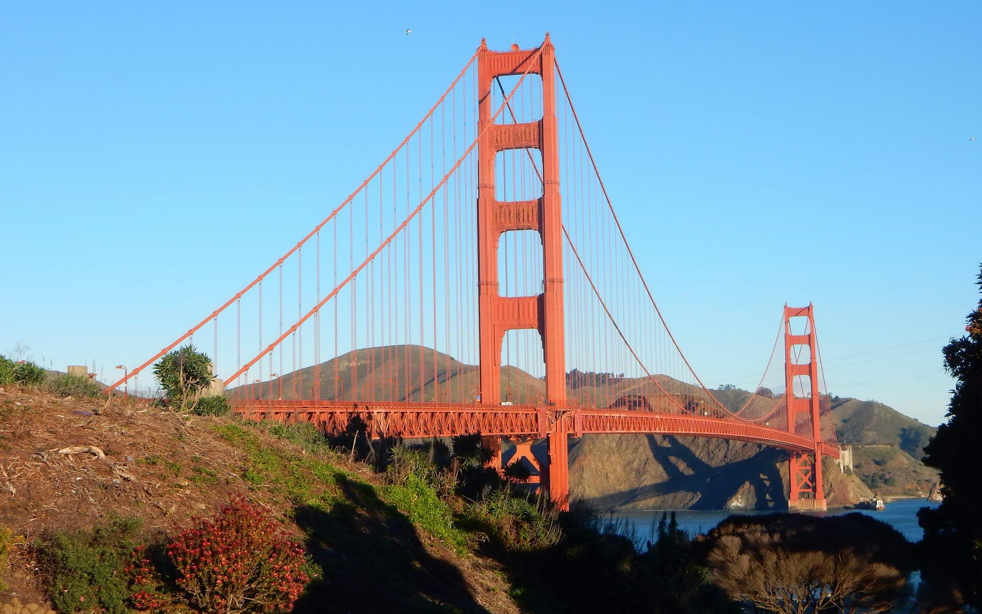 Red suspension bridge over water, with hills and clear blue sky. Sunny.