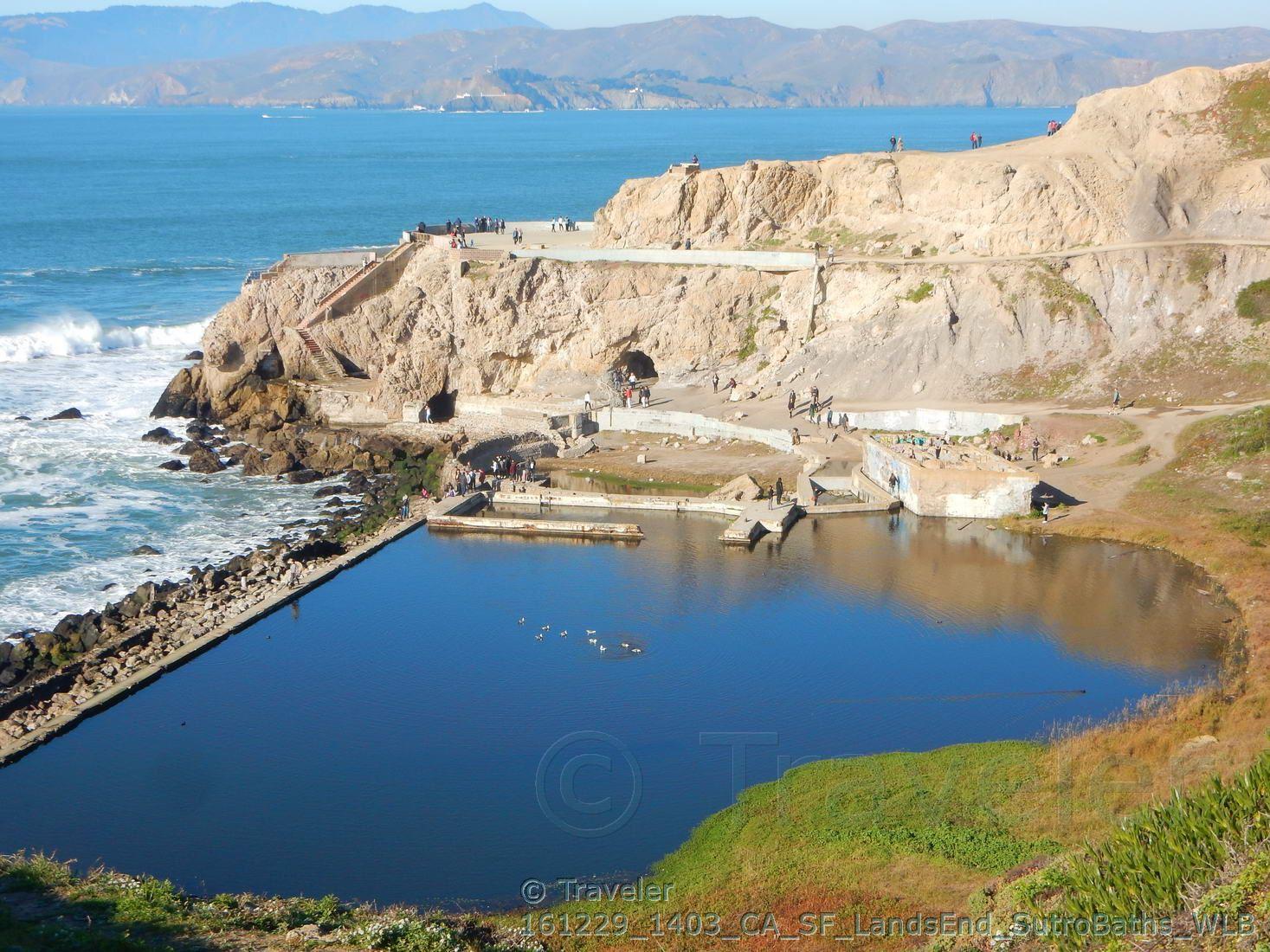 Concrete pools built into a rocky coastline with people, ocean, and distant mountains.