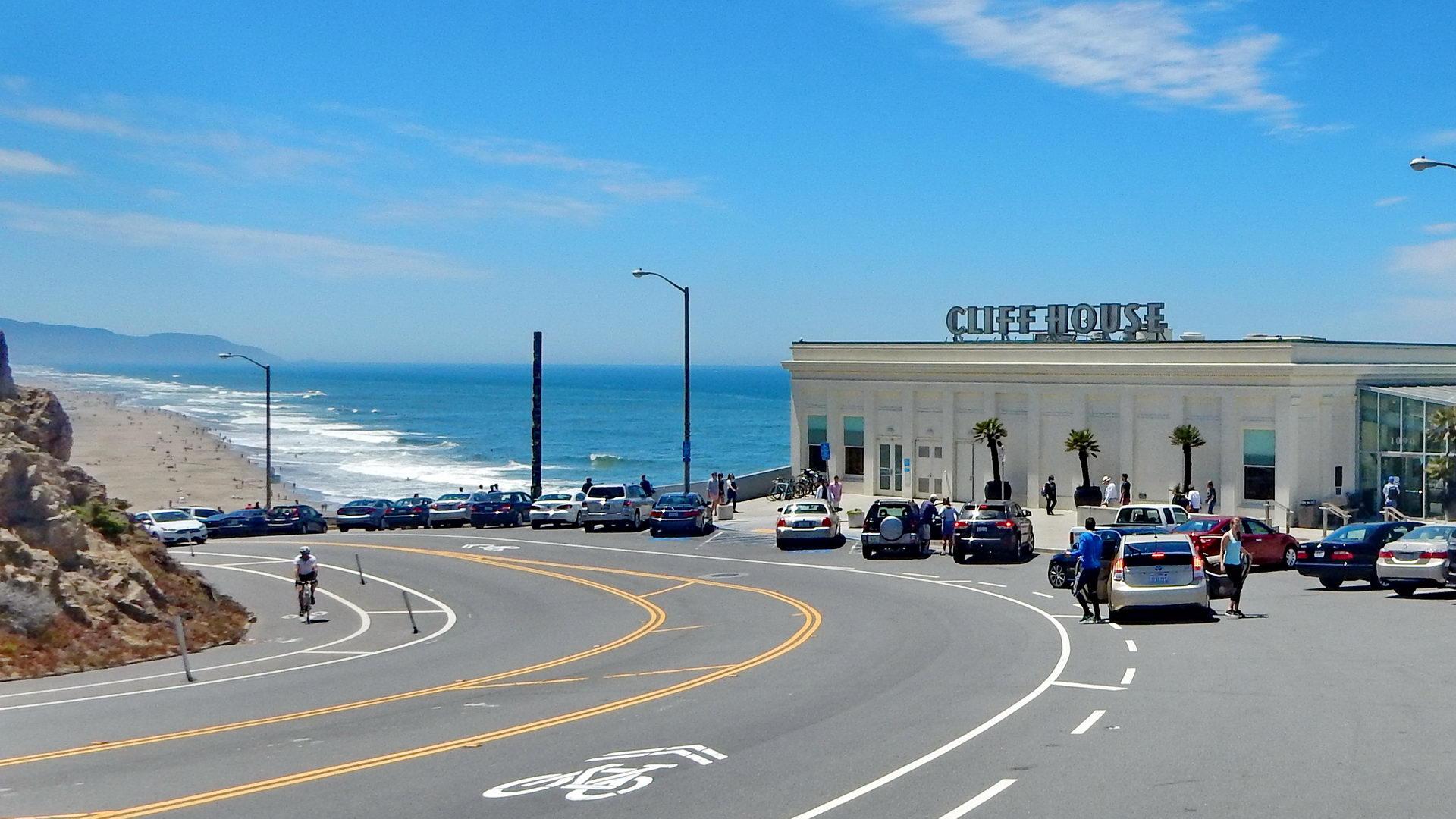 Building next to a curved road, sandy beach, and ocean on a sunny day.