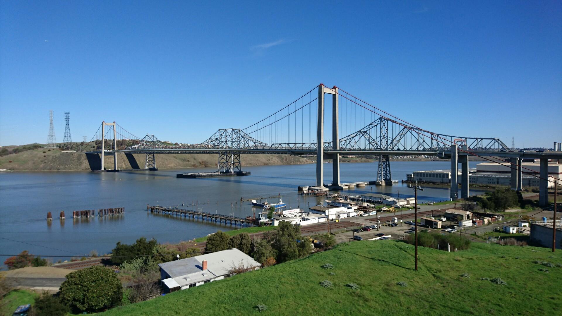 Large suspension bridge over a wide river with hills under bright sun.