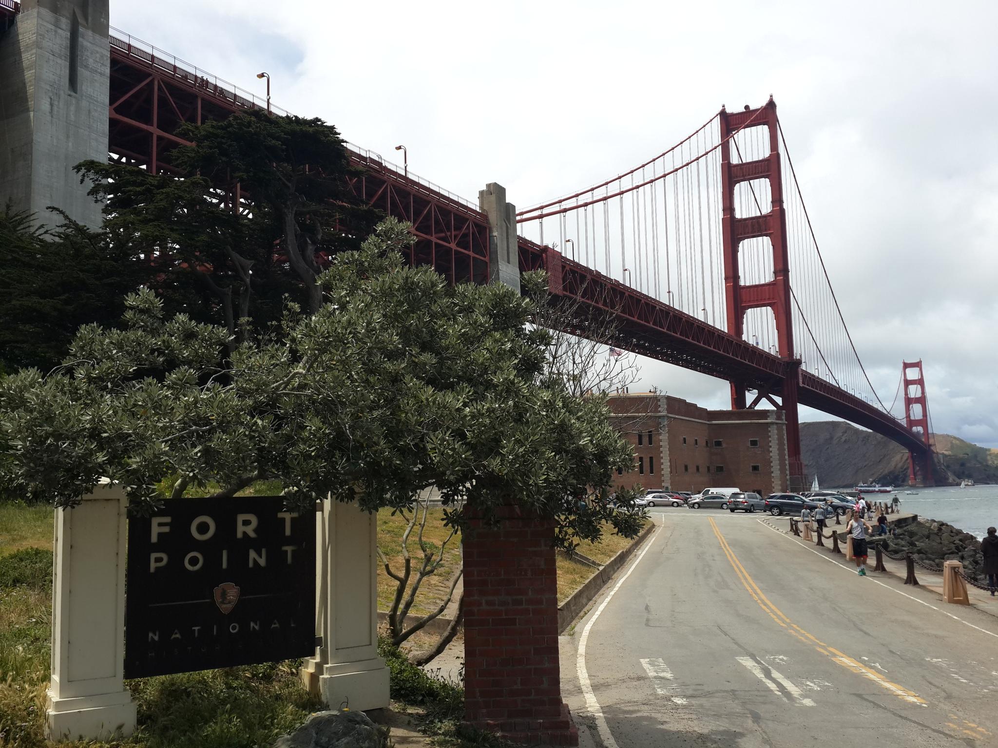 Red suspension bridge from below, with a historic building and road. Overcast.