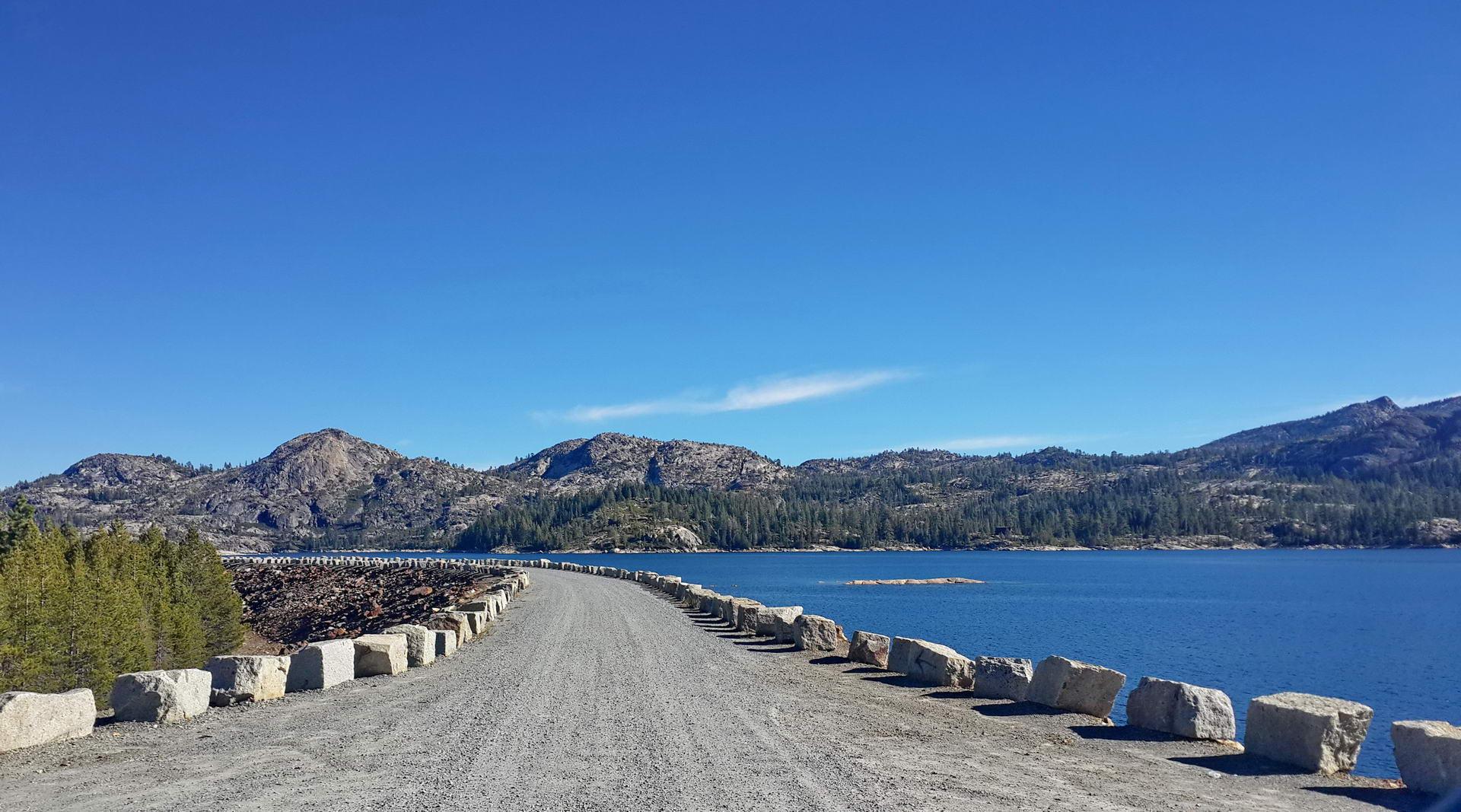 Gravel road on a dam wall beside a large blue lake and mountains.