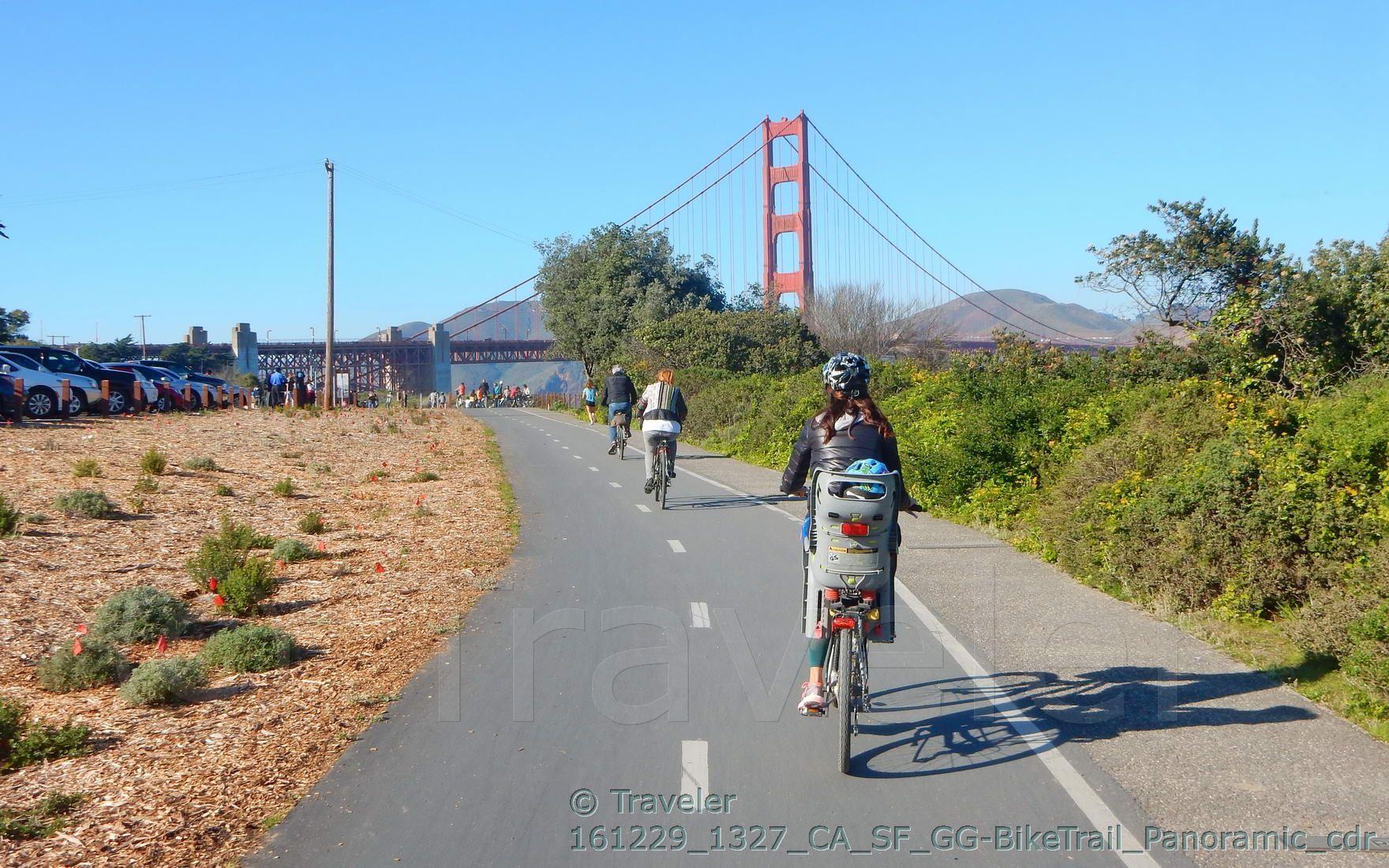 Cyclists ride on a paved path towards a large red suspension bridge on a sunny day.