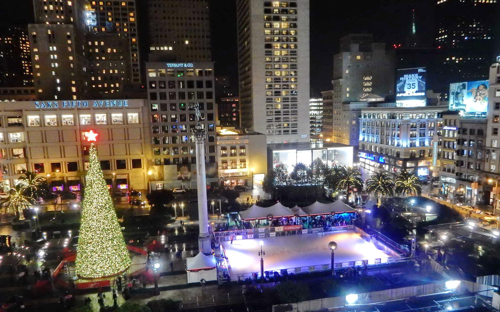 Nighttime city square with a large Christmas tree and ice skating rink.