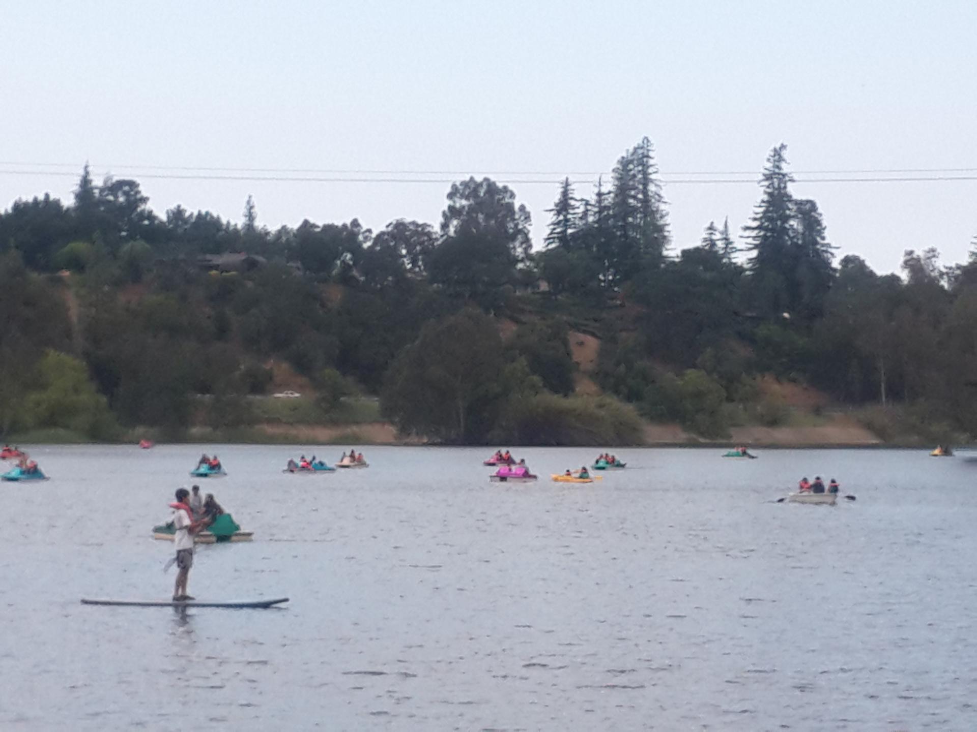 Many people paddleboarding and boating on a lake under an overcast sky.