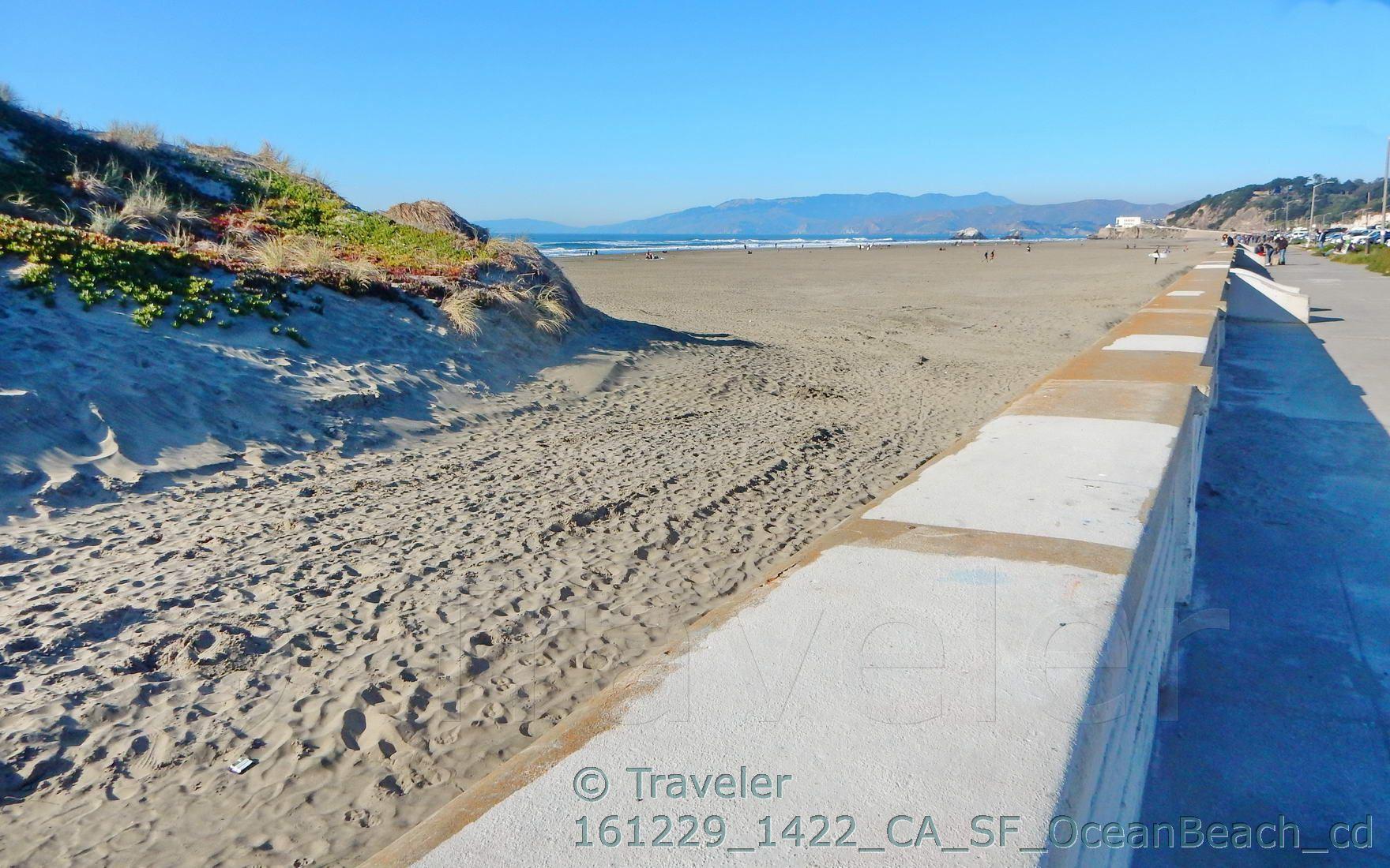 A vast sandy beach next to a concrete wall, with ocean and distant hills.