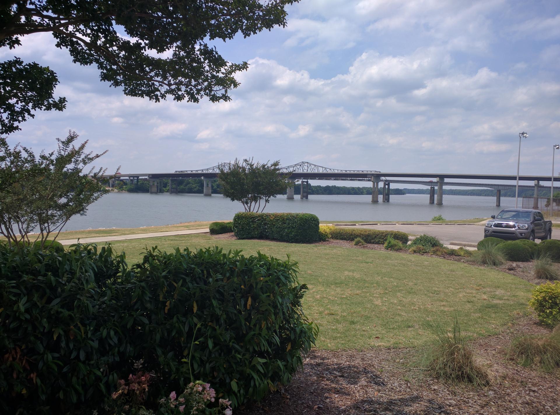 A large bridge spans water, with green foliage in the foreground under a cloudy sky.