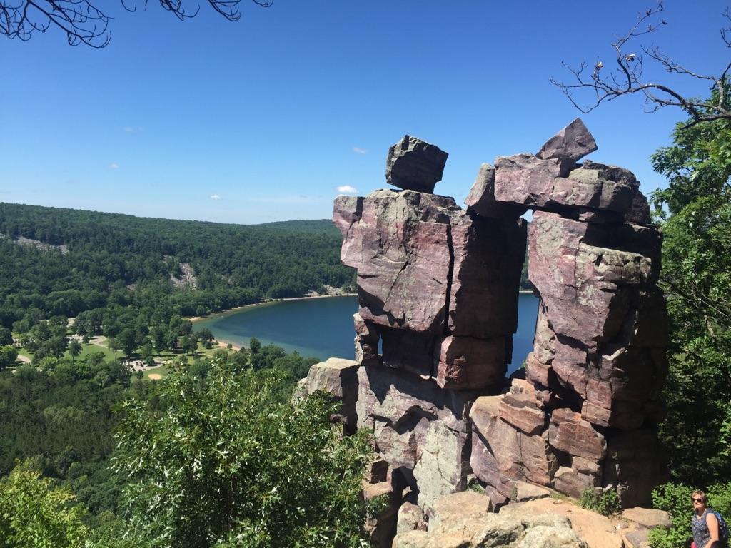 Large rock formation with an arch overlooking a lake and forest, sunny day.