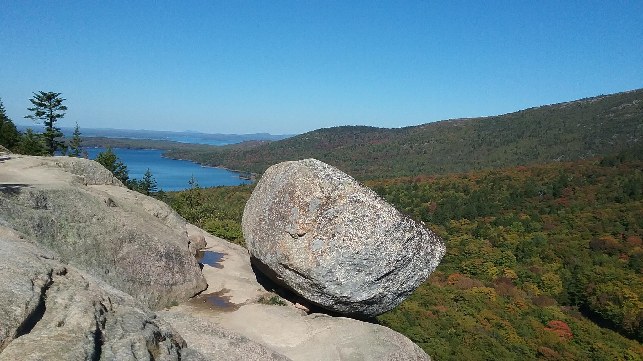 Bright sunny day, large boulder on a rocky cliff overlooking a lake and green hills.