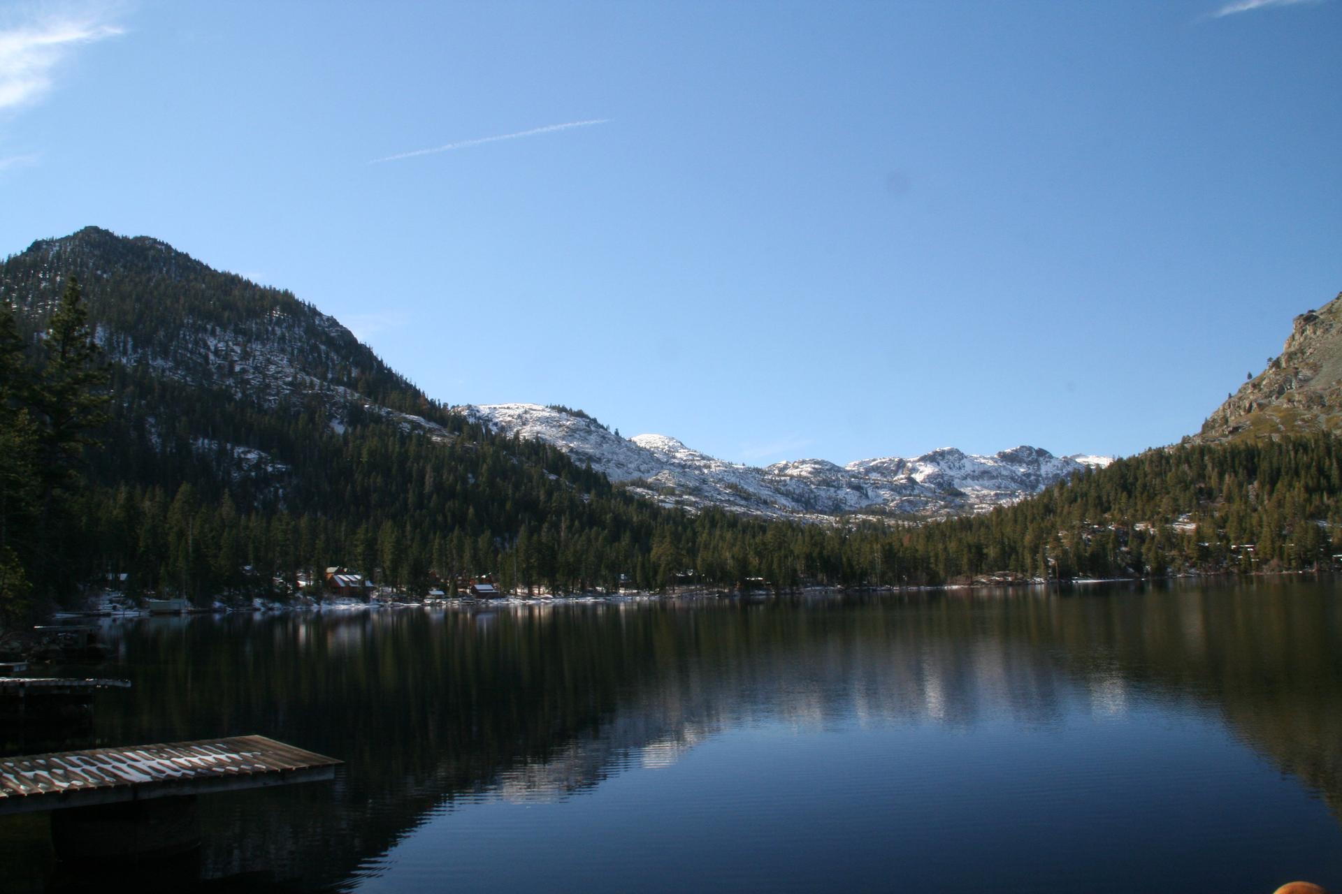 A tranquil lake reflects snow-dusted mountains and pine trees under a bright blue sky.