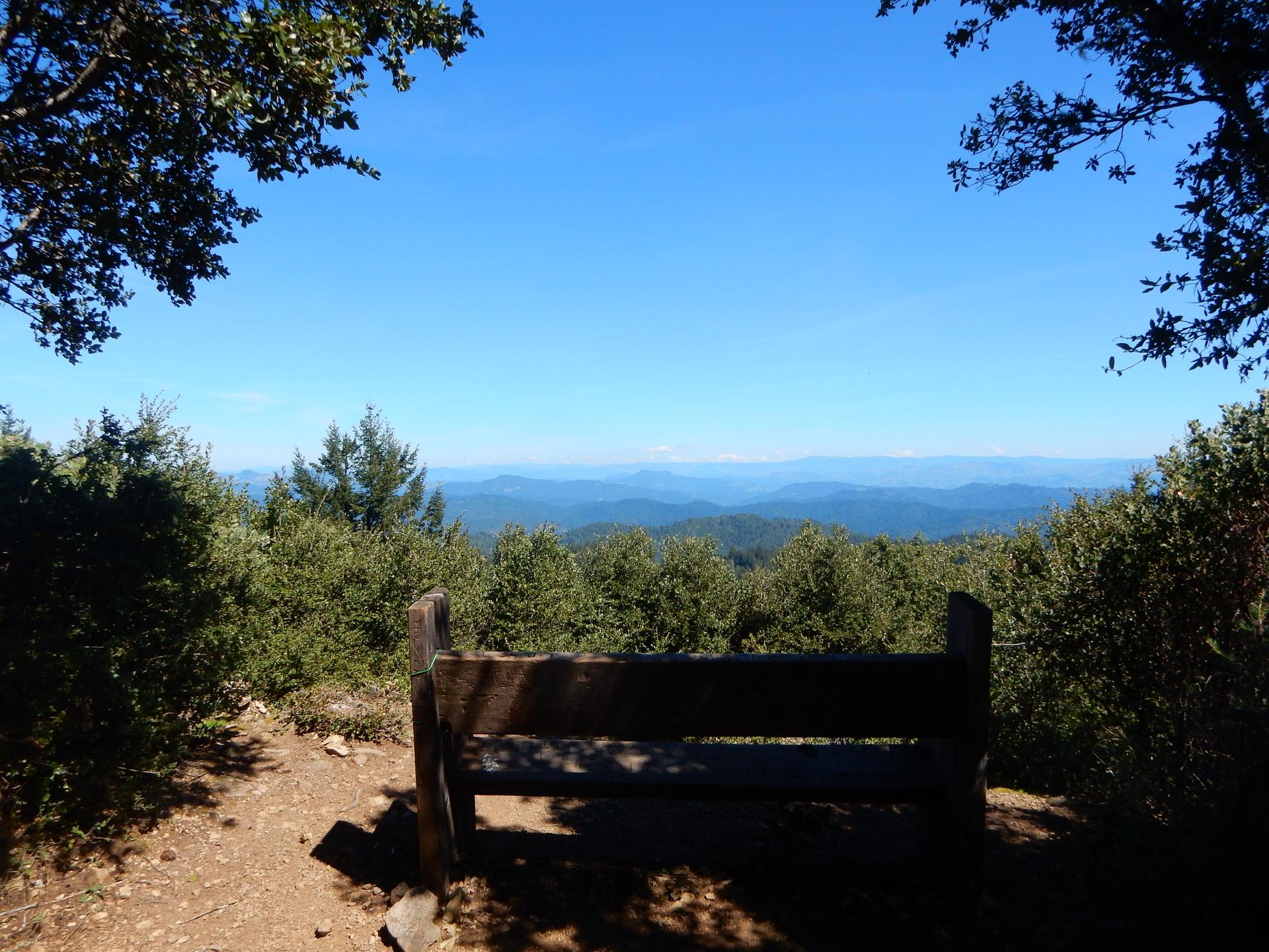 Wooden bench overlooking a vast mountain range under a clear blue sky.