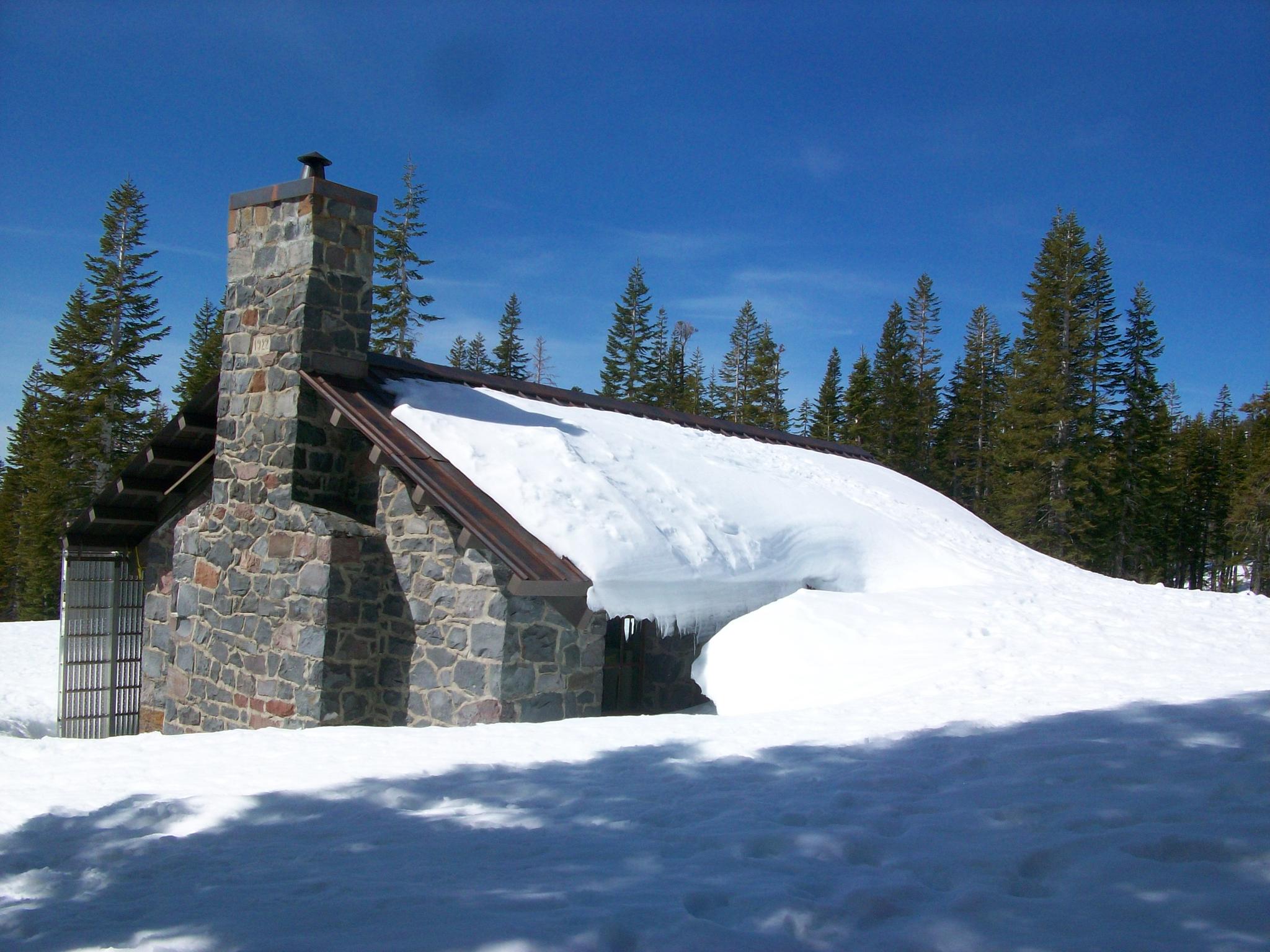 Stone building with a chimney, heavily covered in deep snow under a blue sky.