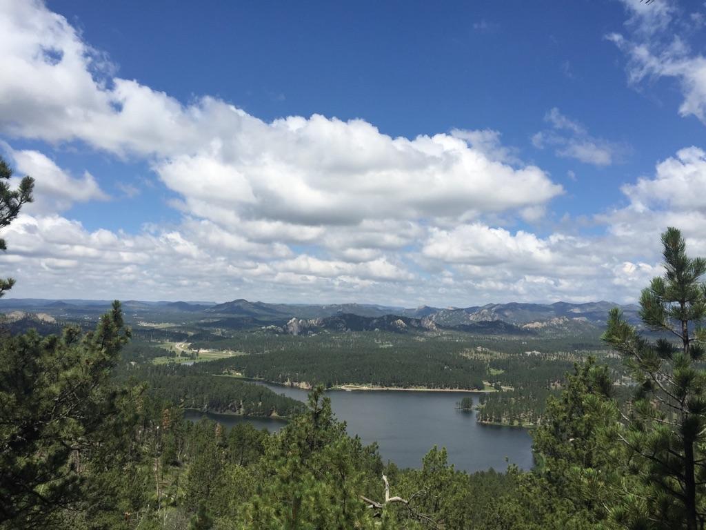 Panoramic view of a lake, dense forests, and distant mountains under a cloudy blue sky.