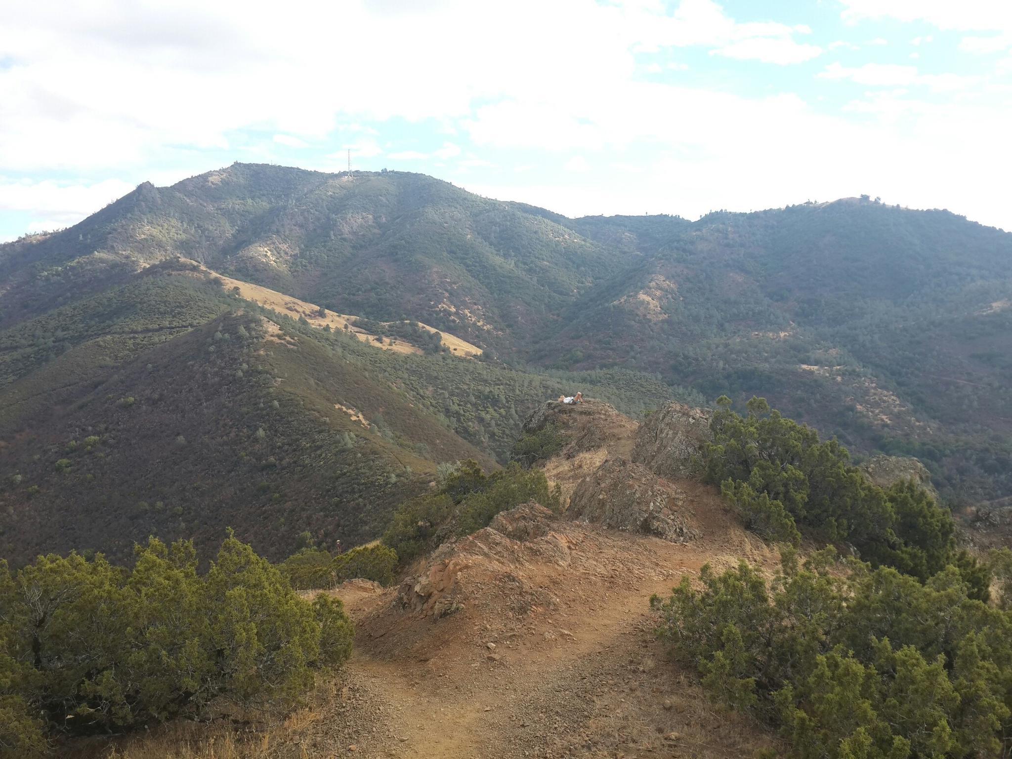 Rocky, dirt trail leading to distant, forested mountains under a cloudy sky.