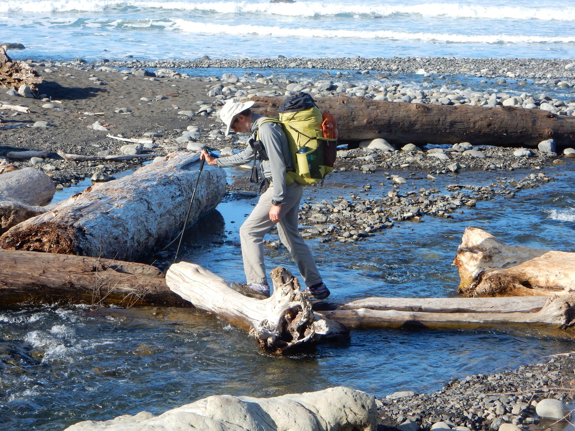 Hiker with backpack crossing a stream on logs on a sunny beach.