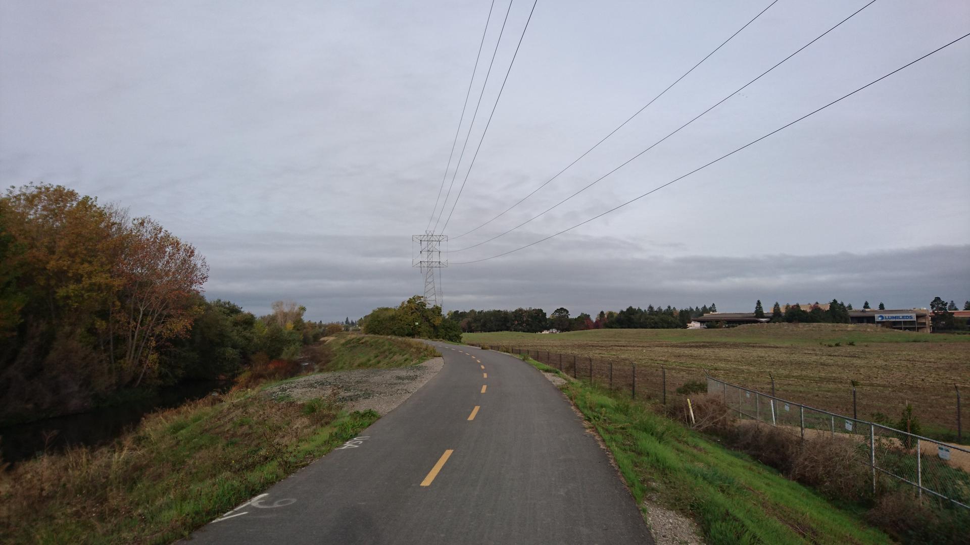Paved path under power lines, with trees and fields on an overcast day.