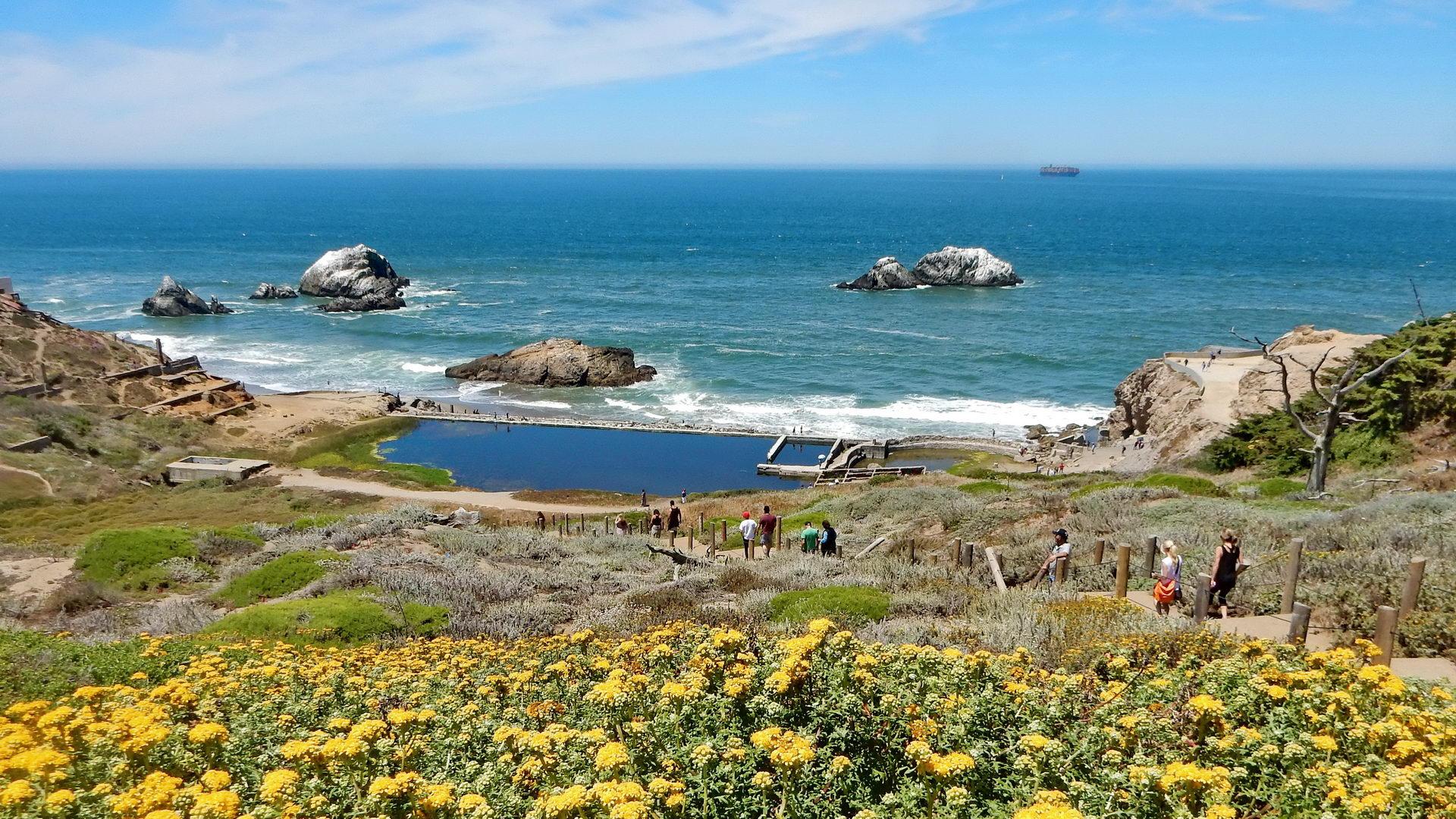 Sunny coastal landscape with ocean, rocky outcrops, old pools, and people.