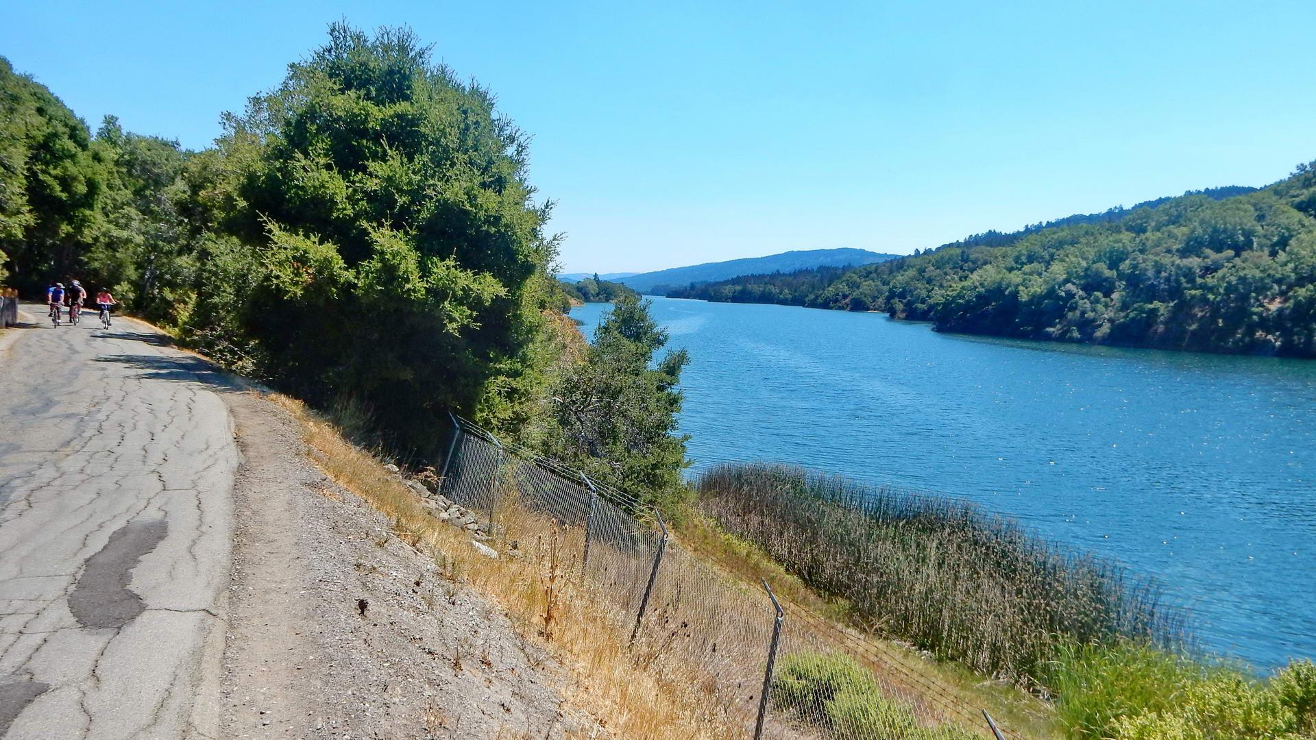 Cyclists on a paved road beside a wide blue river on a sunny day.