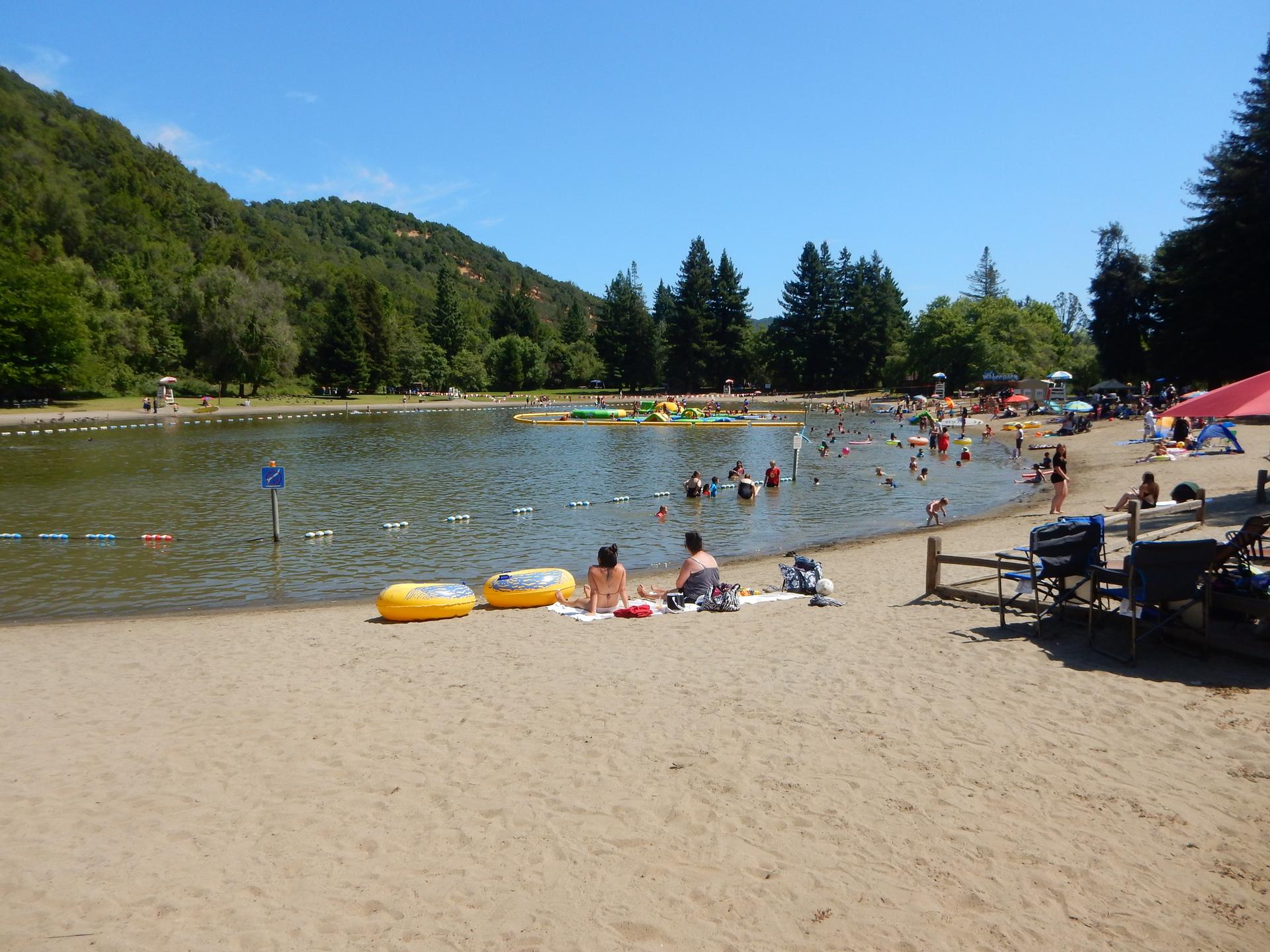 Crowded sandy beach with people swimming and relaxing by a sunny lake.