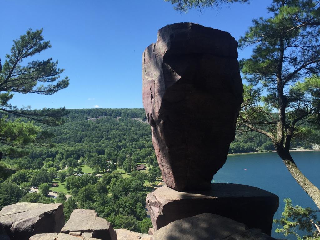Large rock formation on a cliff overlooking a lake and forest under blue sky.