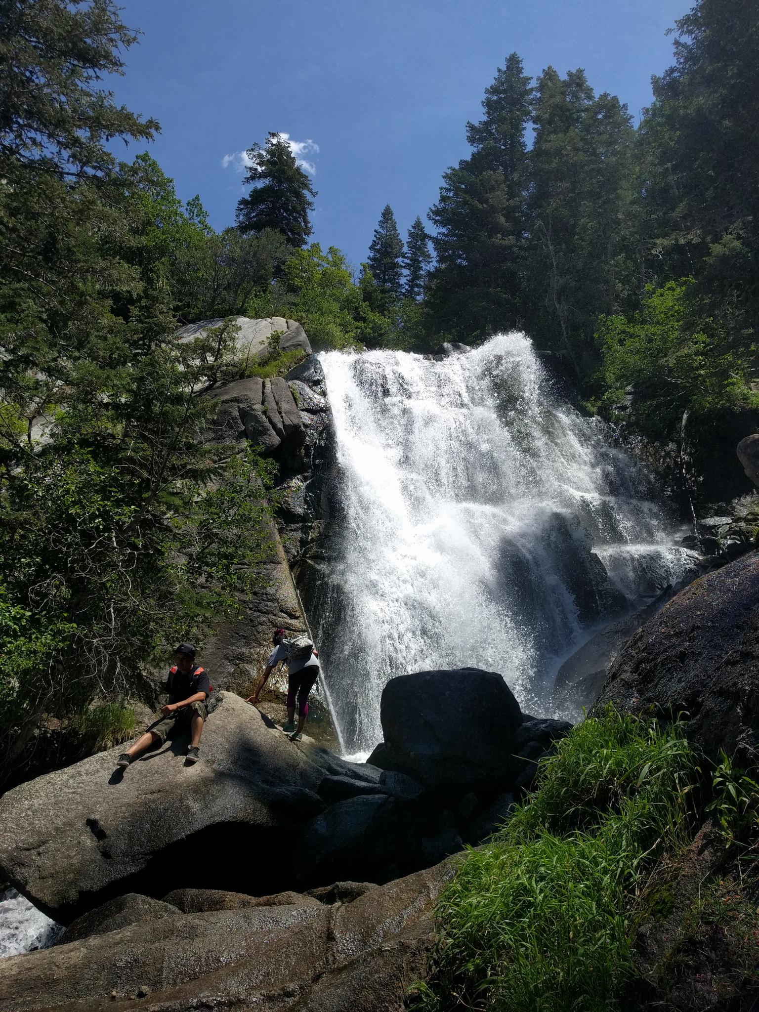 Two people near a powerful waterfall in a sunny, rocky forest.