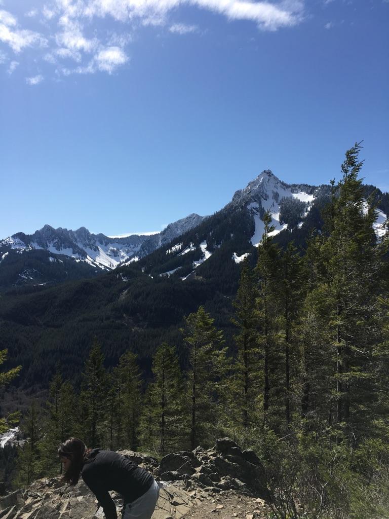 Person on rocky outcrop overlooking vast forested mountains with snow patches.
