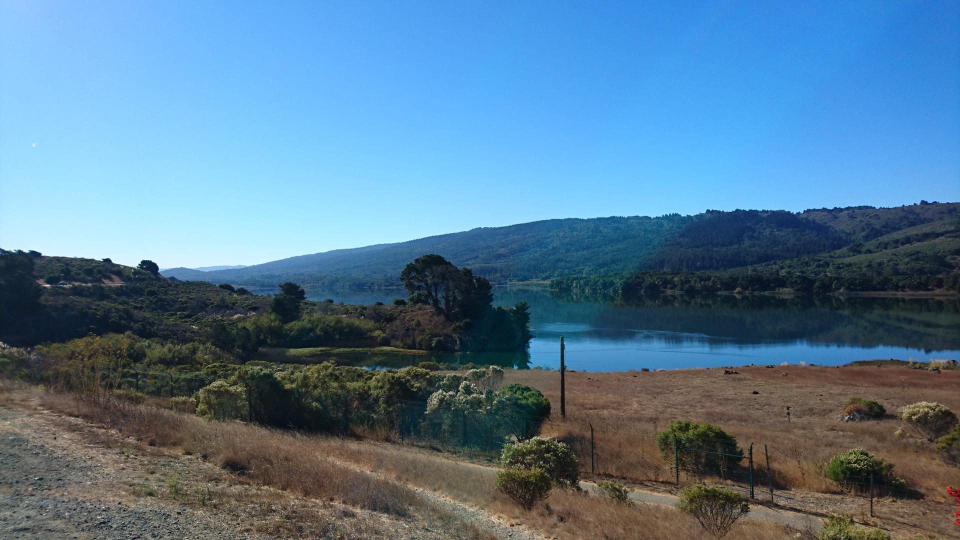 Expansive blue lake surrounded by dry grass and distant green hills under clear sky.