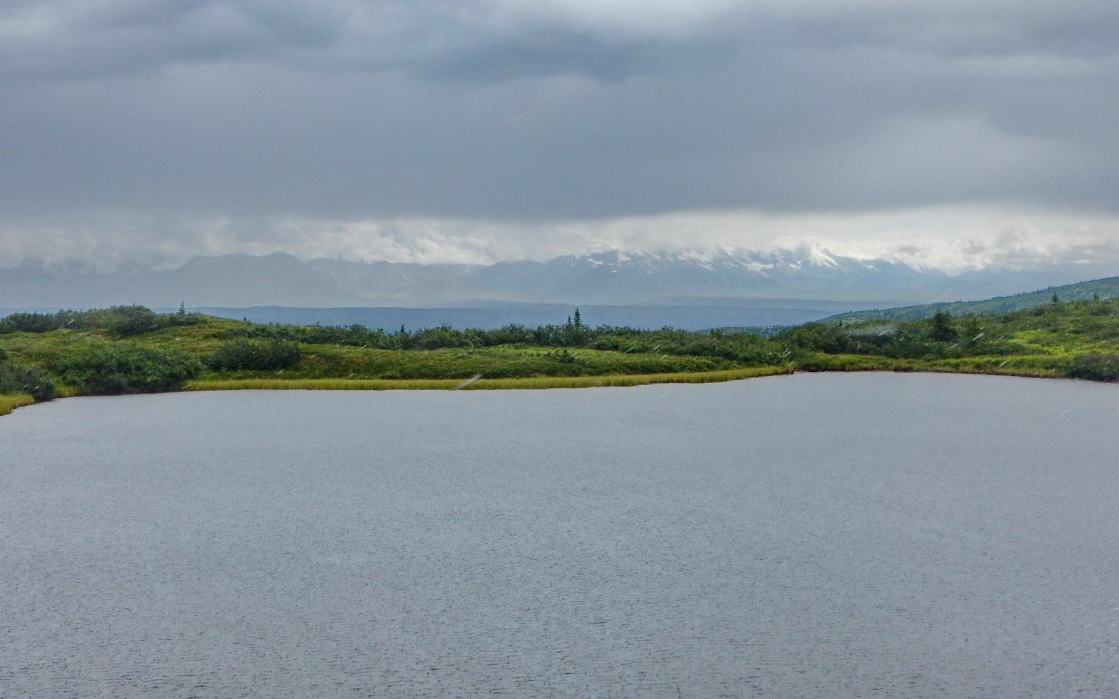 Calm lake with green shores under an overcast sky, distant snowy mountains.