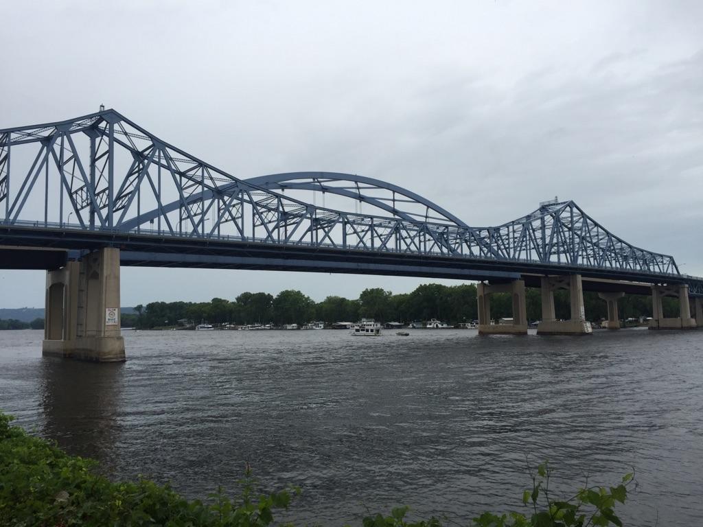 Large blue truss bridge over a wide river with boats, under overcast sky.