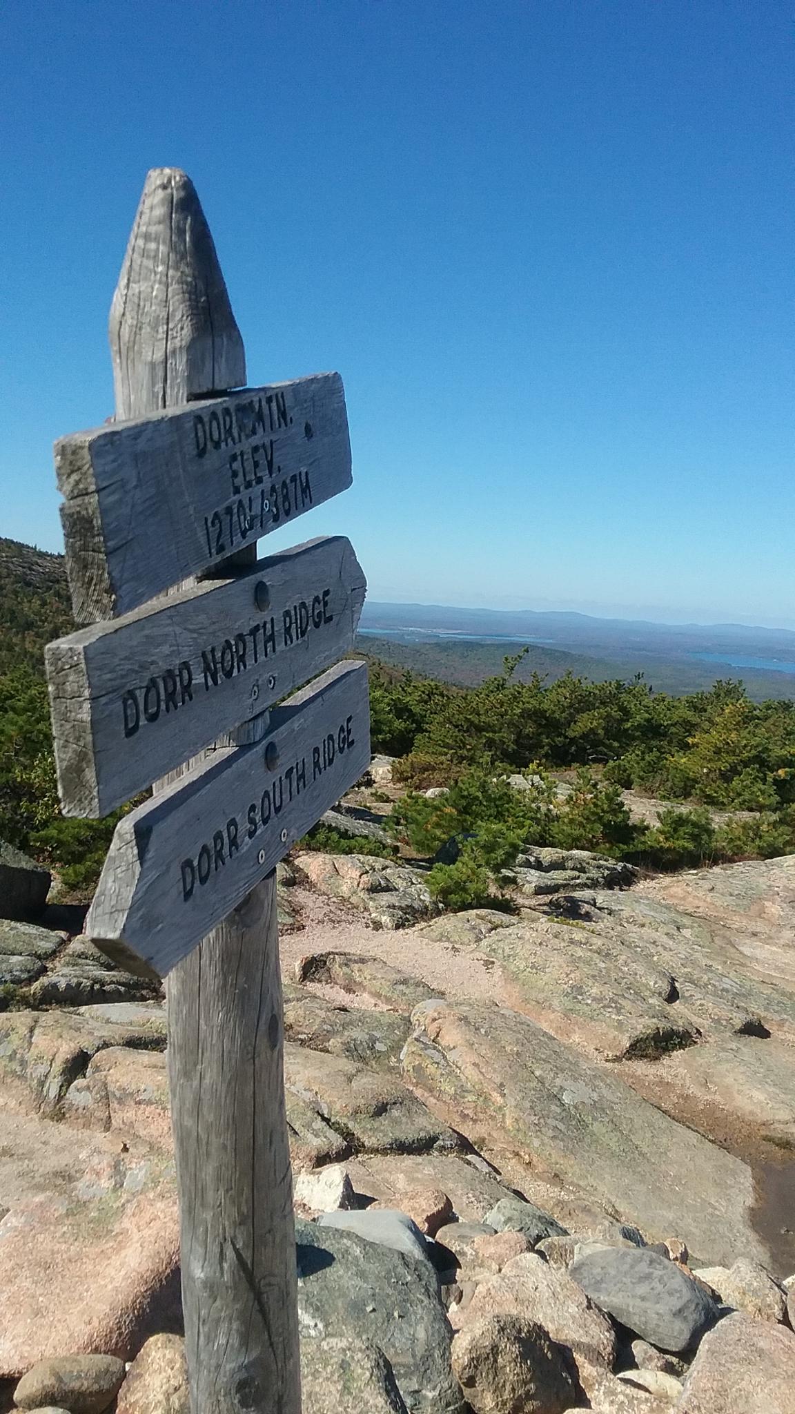 Wooden trail sign on a rocky peak with distant forested hills.