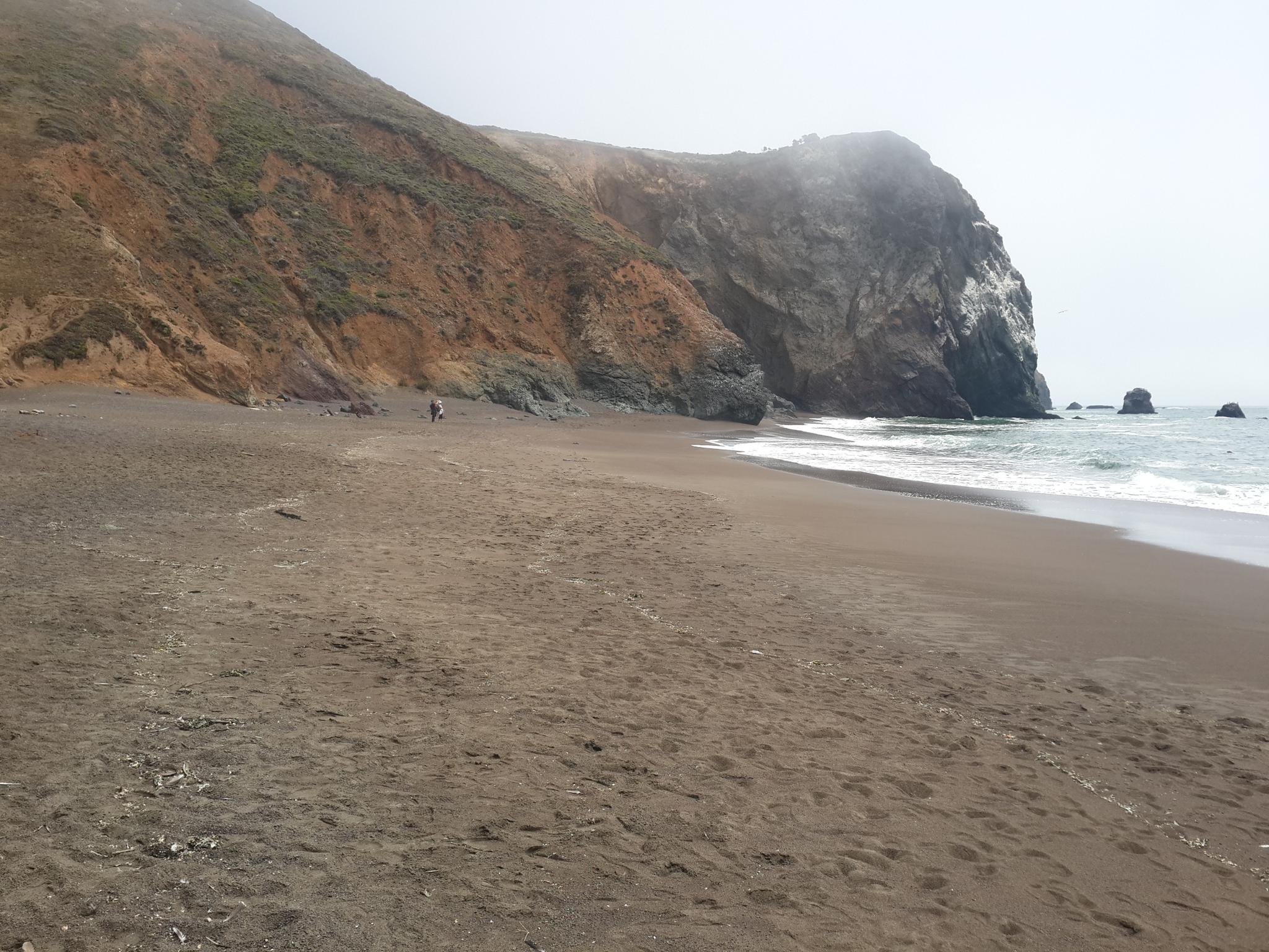 Dark sand beach with large cliffs, ocean waves, and a distant person under overcast sky.